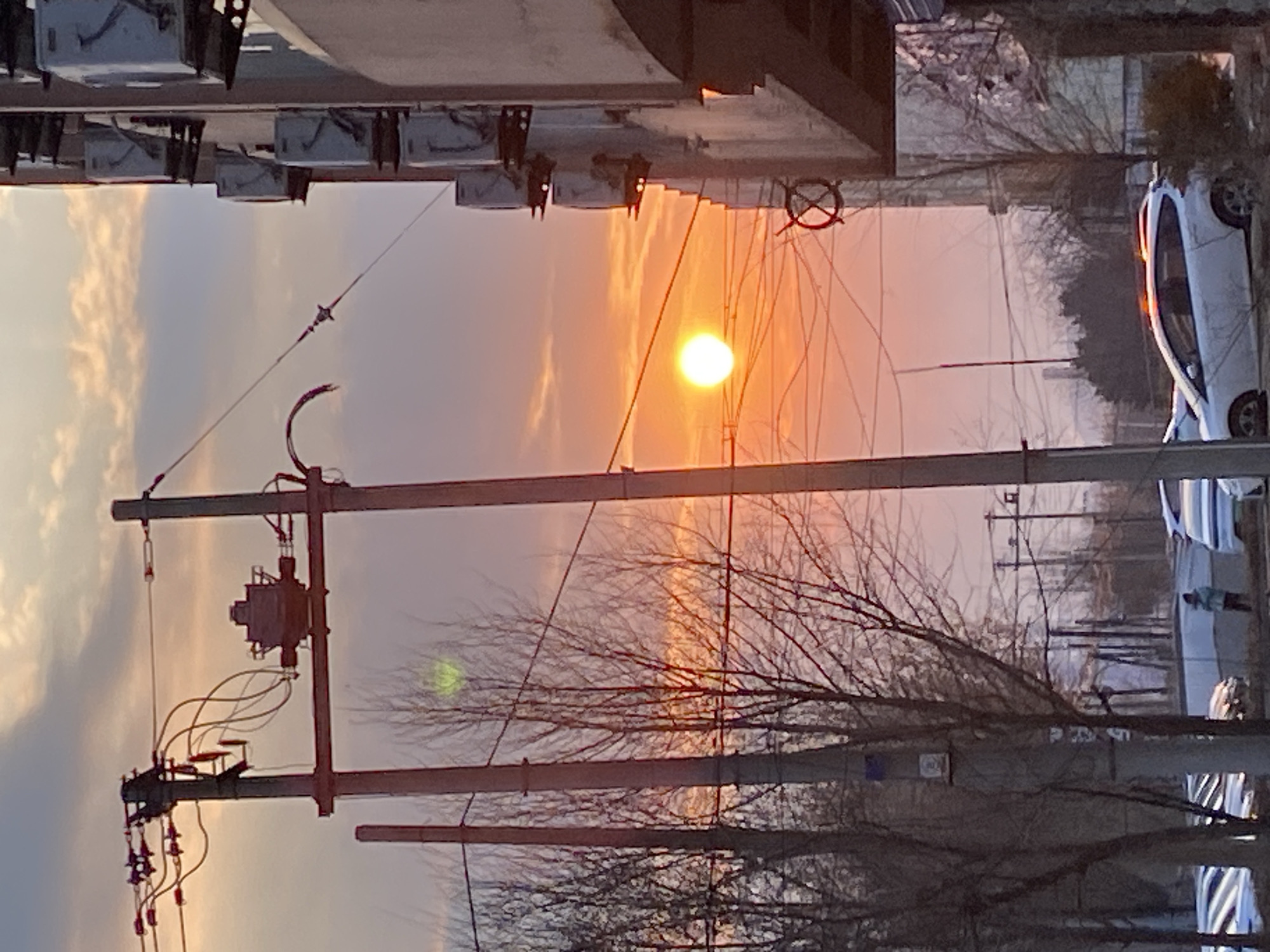 The image captures a sunset in Zhongtun, China. Utility poles with transformers are visible in the foreground. Bare tree branches intersect the view. A white car is parked on the right side of the frame. A building is partially visible at the top left. The sun is positioned in the center of the image, radiating a warm orange glow across the sky.