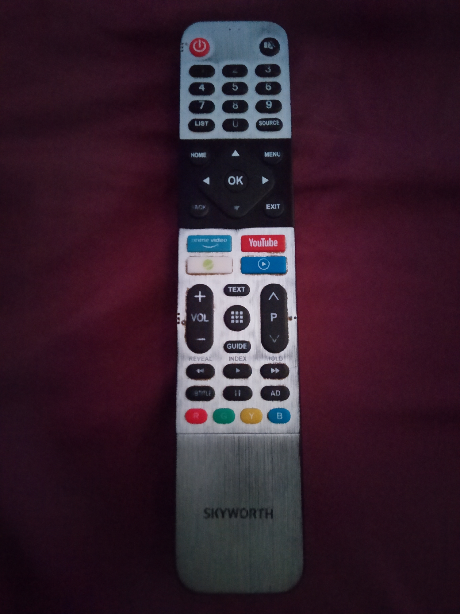 A black and silver TV remote is lying horizontally on a dark red, textured surface. The remote is oriented vertically in the image. The top of the remote has a red power button. Below that are numerical buttons from 0 to 9, along with "LIST" and "SOURCE" buttons. Further down, there are navigation buttons for "HOME," "MENU," "OK," and "EXIT." Below these are dedicated buttons for "prime video" and "YouTube," as well as a play/pause button. The volume controls and channel up/down buttons are present, along with "TEXT," "GUIDE," and "INDEX" buttons. At the bottom are four colored buttons (red, green, yellow, blue) and a silver end piece with "SKYWORTH" printed on it. The image is well-lit, showing the details of the remote clearly. The red surface appears to be fabric, possibly a tablecloth or blanket, with some subtle wrinkles. There are no people or any other objects visible in the image. The location in Tujmazy, Russia, is not discernable from the image itself.