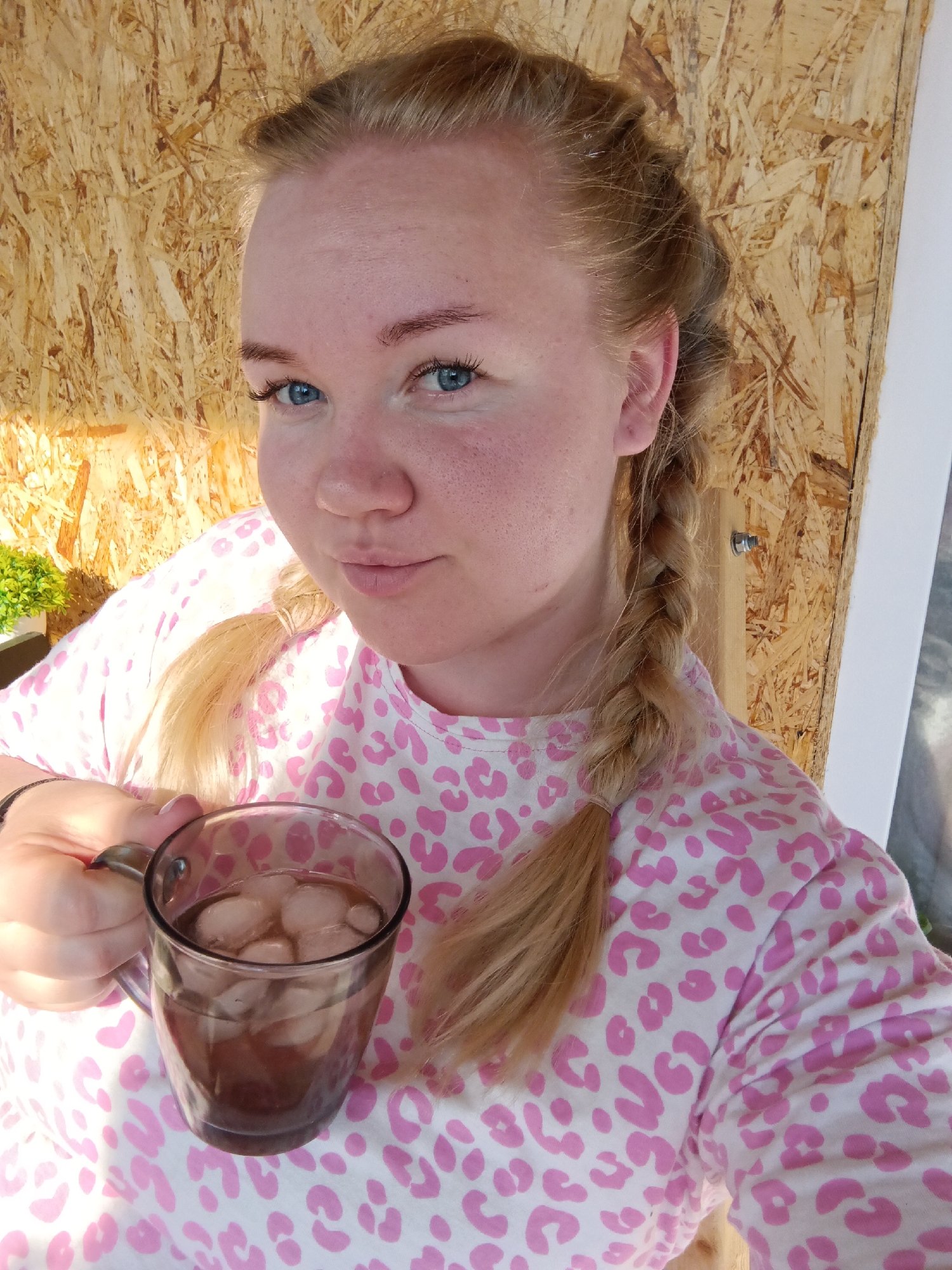 A young woman with light blonde hair styled in pigtail braids holds a glass mug filled with ice and a dark liquid. She is wearing a white shirt with pink leopard print and is positioned in front of a wall constructed from oriented strand board. A small potted plant is partially visible in the background. The location is not discernible from the image.