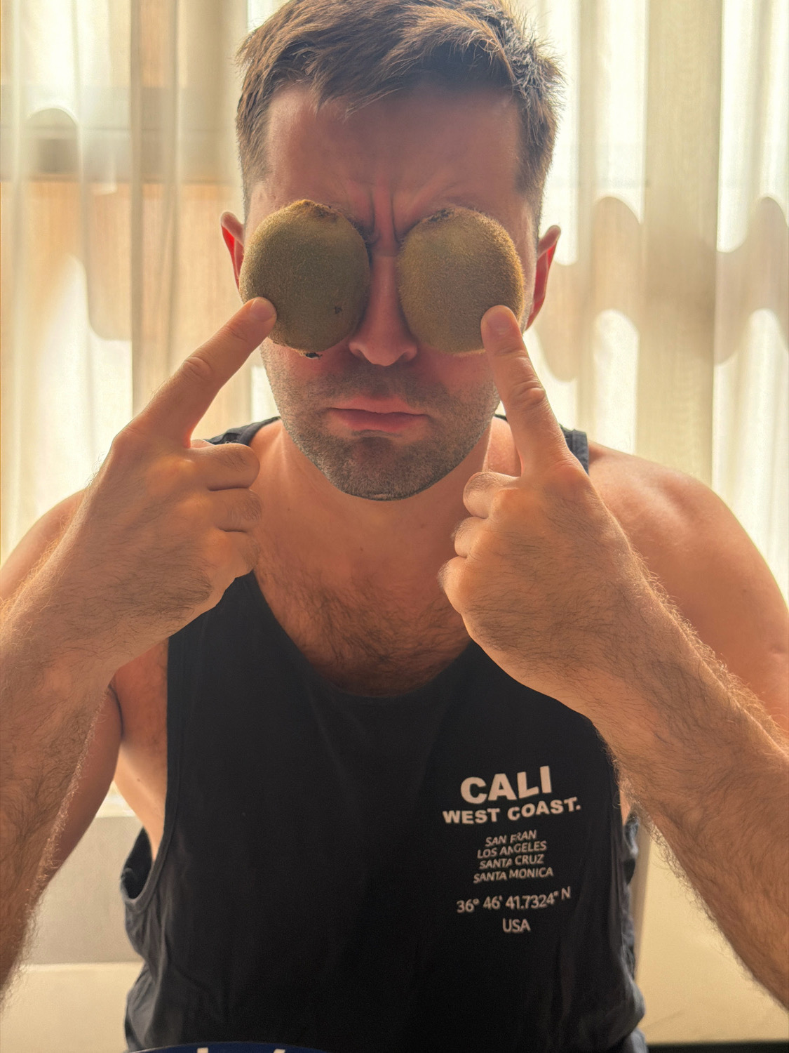 A Caucasian man with short brown hair is holding two kiwis over his eyes, his fingers are visible, and he is looking at the camera with a serious expression. He is wearing a black tank top with "CALI WEST COAST" printed on it. The background is a blurred curtain. The image does not depict any elements of Verona, Italy.