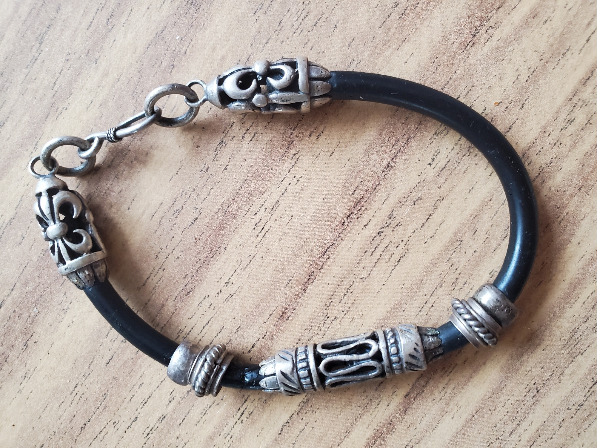 The image shows a black rubber bracelet with silver-colored decorative elements on a light brown wooden surface. The bracelet is curved, forming an oval shape. It has several ornate silver beads and spacers. One end of the bracelet features a clasp mechanism composed of multiple interconnected silver rings. The wooden surface has a visible grain pattern and appears to be a tabletop or similar flat surface. There are no people or animals present in the image, and no specific geographical landmarks are identifiable to indicate its location in Tiraspol, Moldova.