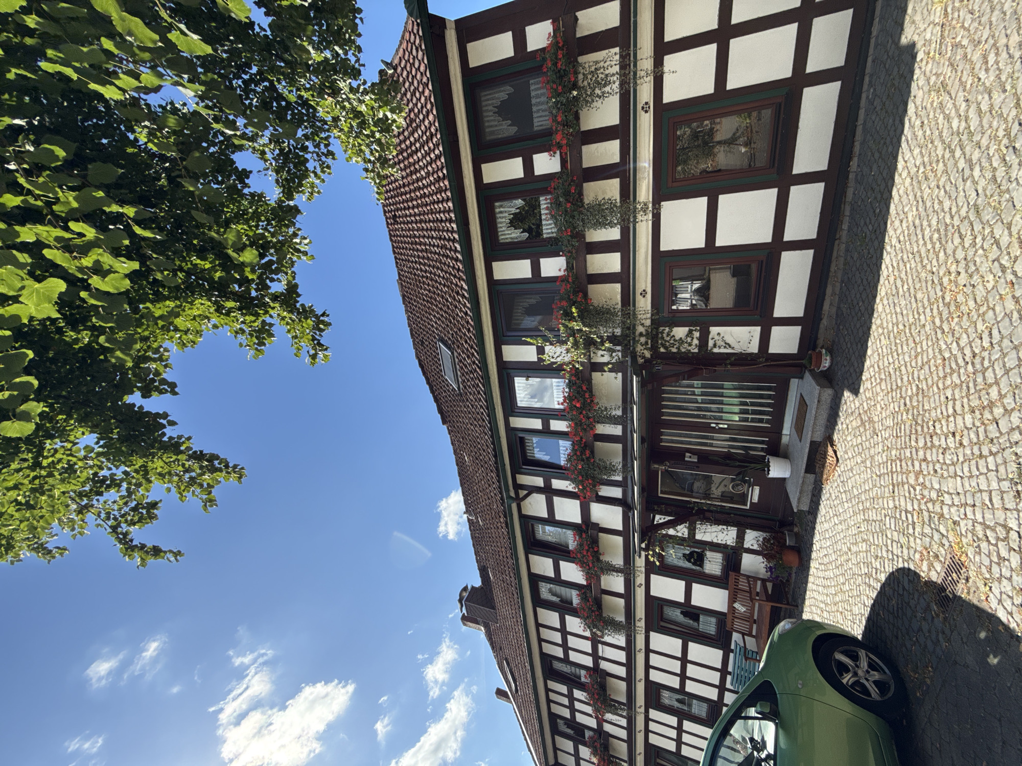 A half-timbered building with a tiled roof stands in Walkenried, Germany, under a bright blue sky with scattered white clouds. Green foliage from a tree hangs over the top of the image. The building's facade features white infill between dark brown timbers, with numerous windows adorned with red flowers in window boxes and cascading greenery. A dark brown double door with vertical panes serves as the entrance, approached by stone steps. To the left of the entrance, a wooden bench and a green car are parked on a cobblestone driveway. The overall scene depicts a charming, well-maintained residential or commercial property in a European town.