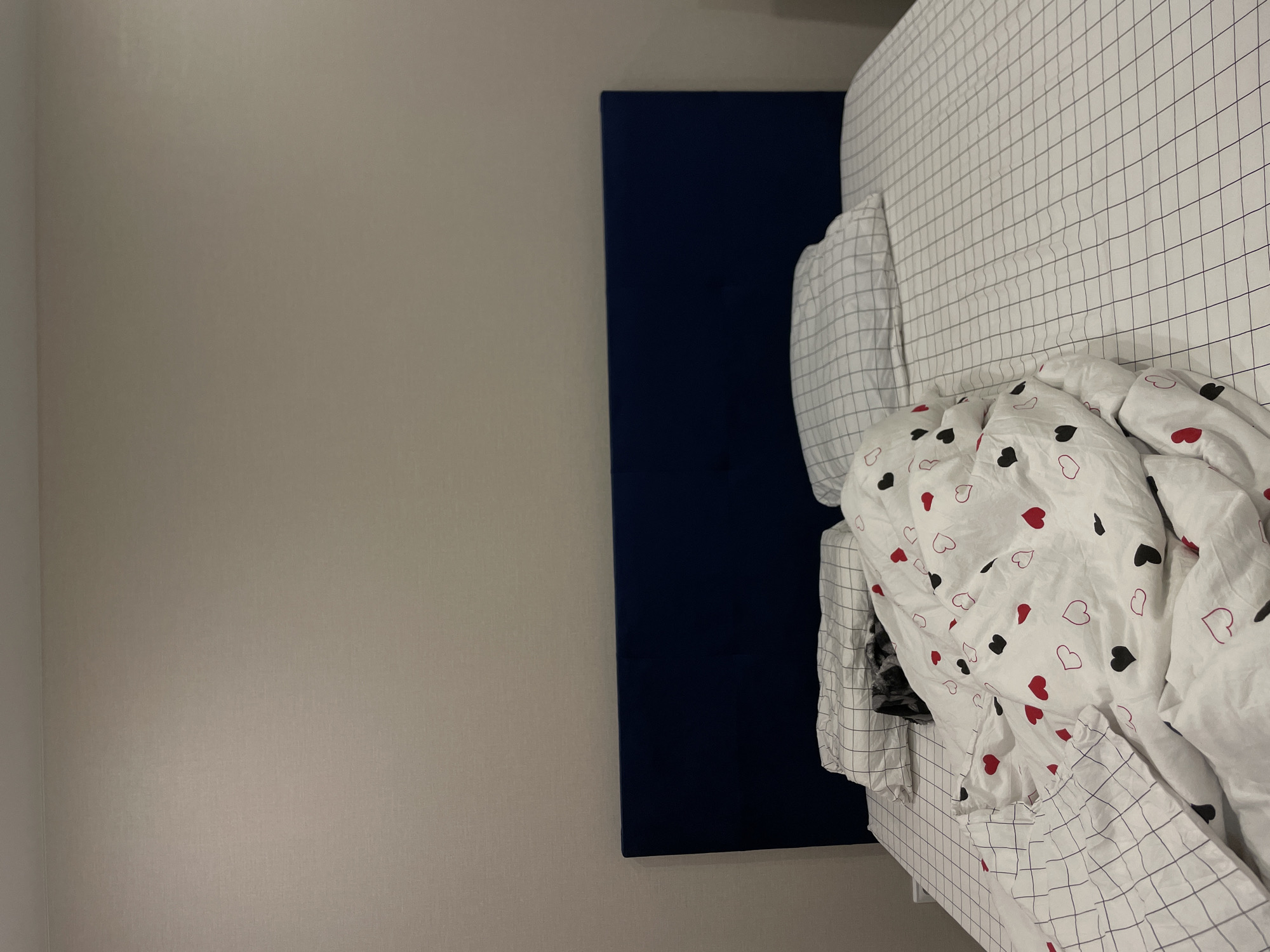 The image depicts an indoor scene, likely a bedroom, with a bed prominently featured on the right side of the frame. The bed is adorned with white bedding; the fitted sheet and pillowcases display a minimalist grey grid pattern on a white background. A rumpled duvet cover, also white, features an assortment of red and black solid hearts, along with red outlined hearts. Behind the bed is a dark blue headboard. To the left of the bed, a plain light beige or off-white wall fills the rest of the space, with a subtle corner visible on the far left. The lighting in the room appears soft and even. No specific activity is taking place, and the bed appears unmade. There is no visible text in the image. The general setting suggests a domestic environment in Podshivalovo, Russia, although no specific regional cues are visible in the room itself.