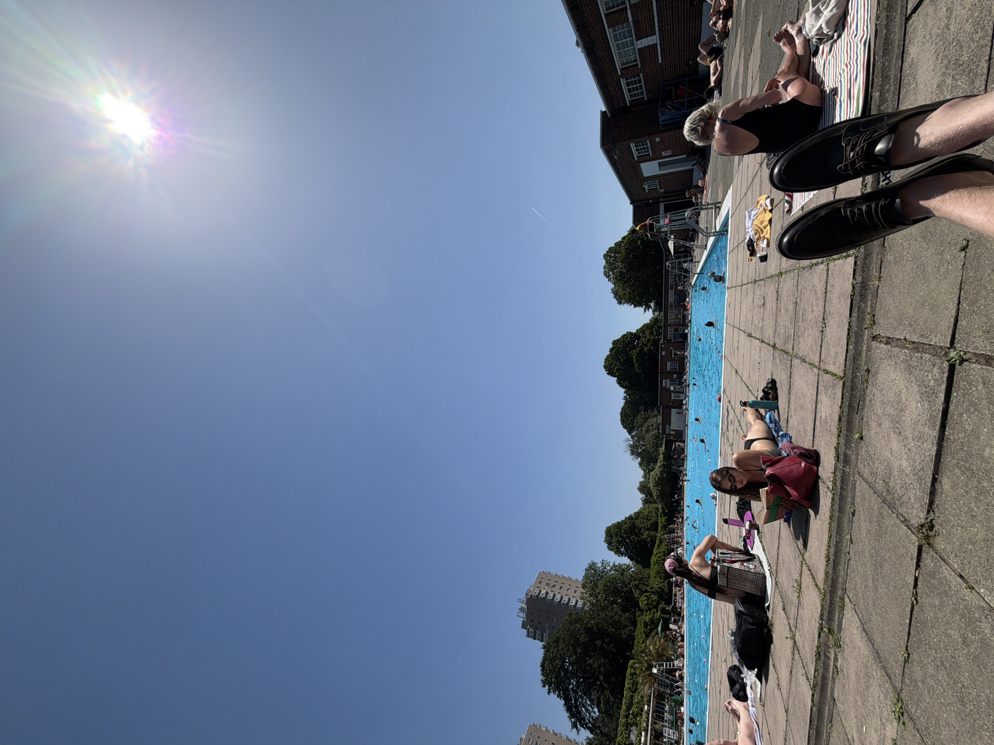 Outdoor scene at a lido in London, UK, on a sunny day. People are sunbathing and swimming in the pool. A brick building borders one side of the pool. Trees and a high-rise building are in the background. In the foreground are two feet with shoes.
