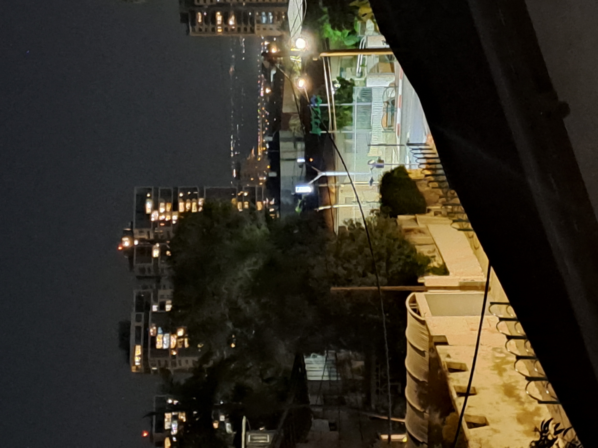 The image shows a nighttime view of a city street in Ramat HaSharon, Israel. In the foreground, a dark, angled roof covers the bottom portion of the frame. Behind it, a light-colored wall runs horizontally, featuring arched structures on top and decorative fencing along its base. Further into the scene, a road is visible with traffic lights, streetlights, and a bench. Beyond the street, the middle ground contains buildings and dense foliage from trees. In the background, a cityscape unfolds with several multi-story buildings illuminated by lights from their windows, suggesting residential or office spaces. The distant horizon shows a faint line of lights, possibly indicating a larger urban area or coastline. The overall scene depicts a quiet urban environment at night, with residential and infrastructure elements visible.