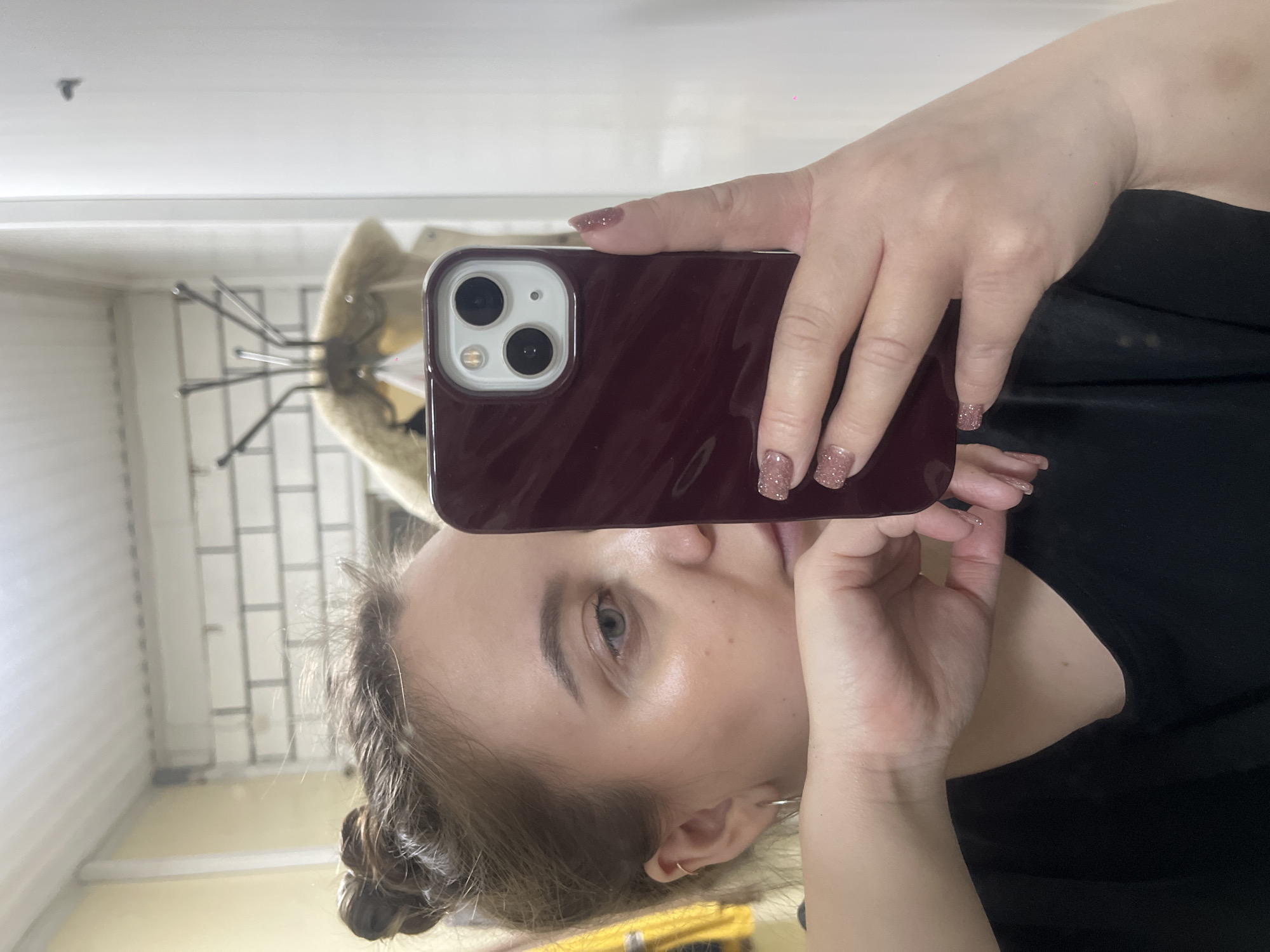 A woman takes a selfie in a room with a reflective surface. She holds a maroon smartphone with a dual-lens camera in her right hand. The phone case is solid in color. Her left hand is near her face. She has fair skin, light eyes, and brown hair styled in a low bun. Her nails are manicured with a glittery polish. She is wearing a black top. In the reflection, a coat stand with a light-colored coat can be seen. The walls appear to be white or light gray, and the room is moderately illuminated. The location is unspecified, and the image does not contain information regarding Novopokrovskaya, Russia.