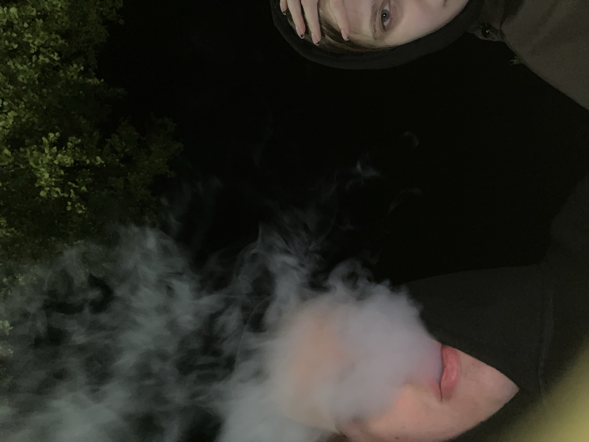 The photograph, taken at night in Красные Баки, Russia, captures a low-angle perspective of two individuals in an outdoor setting. The scene is dominated by darkness, with faint illumination highlighting certain elements.
In the upper part of the frame, the upper half of one person's face is visible, including a light-colored eye and part of their forehead. Their fingers, adorned with light-colored nails with dark tips, rest near their face. This person appears to be wearing a dark hooded garment.
In the lower portion of the image, the chin, pink lips, and part of the cheek of a second person are visible. This individual is actively exhaling a large, dense cloud of white vapor or smoke, which spreads across the lower-left and central areas of the frame.
To the far left of the image, dimly lit green foliage, likely from a tree or bush, is partially visible, confirming an outdoor location. The overall mood is casual and dimly lit, characteristic of a nighttime shot. No text is visible in the image.