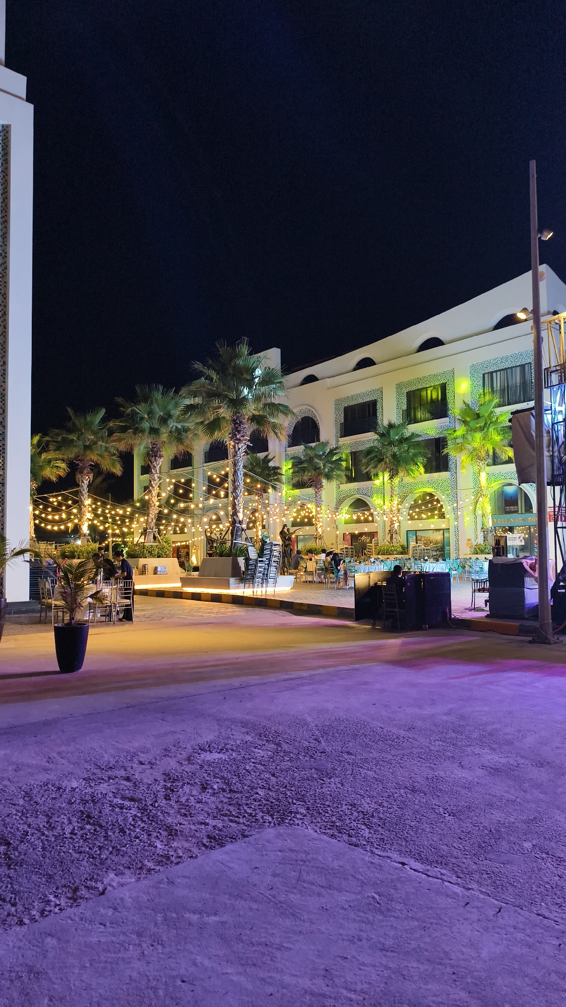 The image depicts an outdoor seating area of a restaurant or establishment at night in the city of Bac Nha Trang Ward, Vietnam. The scene is illuminated by numerous string lights draped between palm trees, casting a warm glow. A multi-story building with arched windows and ornate green tilework serves as the backdrop. Several tables and chairs are set up, with a few patrons seated, suggesting the establishment is open for business. The ground in the foreground appears to be a paved surface with a rough texture, bathed in purple lighting. To the left, a large potted plant is visible. The overall ambiance is festive and inviting, characteristic of a nighttime dining or social venue.