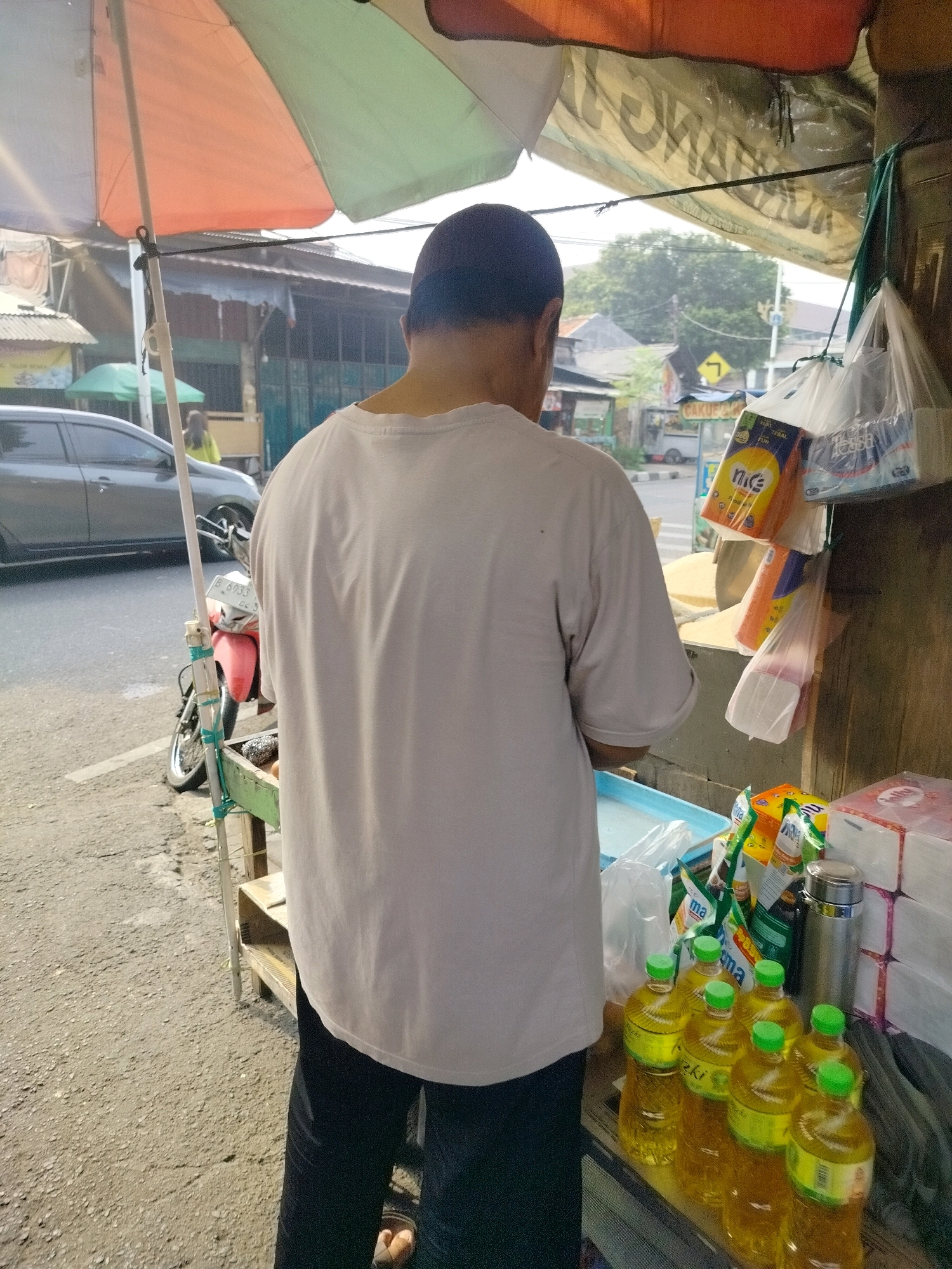The image shows a back view of a person wearing a light-colored t-shirt and dark pants standing near a street vendor stall in East Jakarta, Indonesia. The vendor stall appears to be set up on the side of a busy road, with cars and a motorcycle visible in the background. The stall is covered by an umbrella and displays various items, including bottles of cooking oil, packaged goods like tissues, and what appears to be rice or grains in sacks. The overall scene suggests a typical street-side business operation in an urban environment.