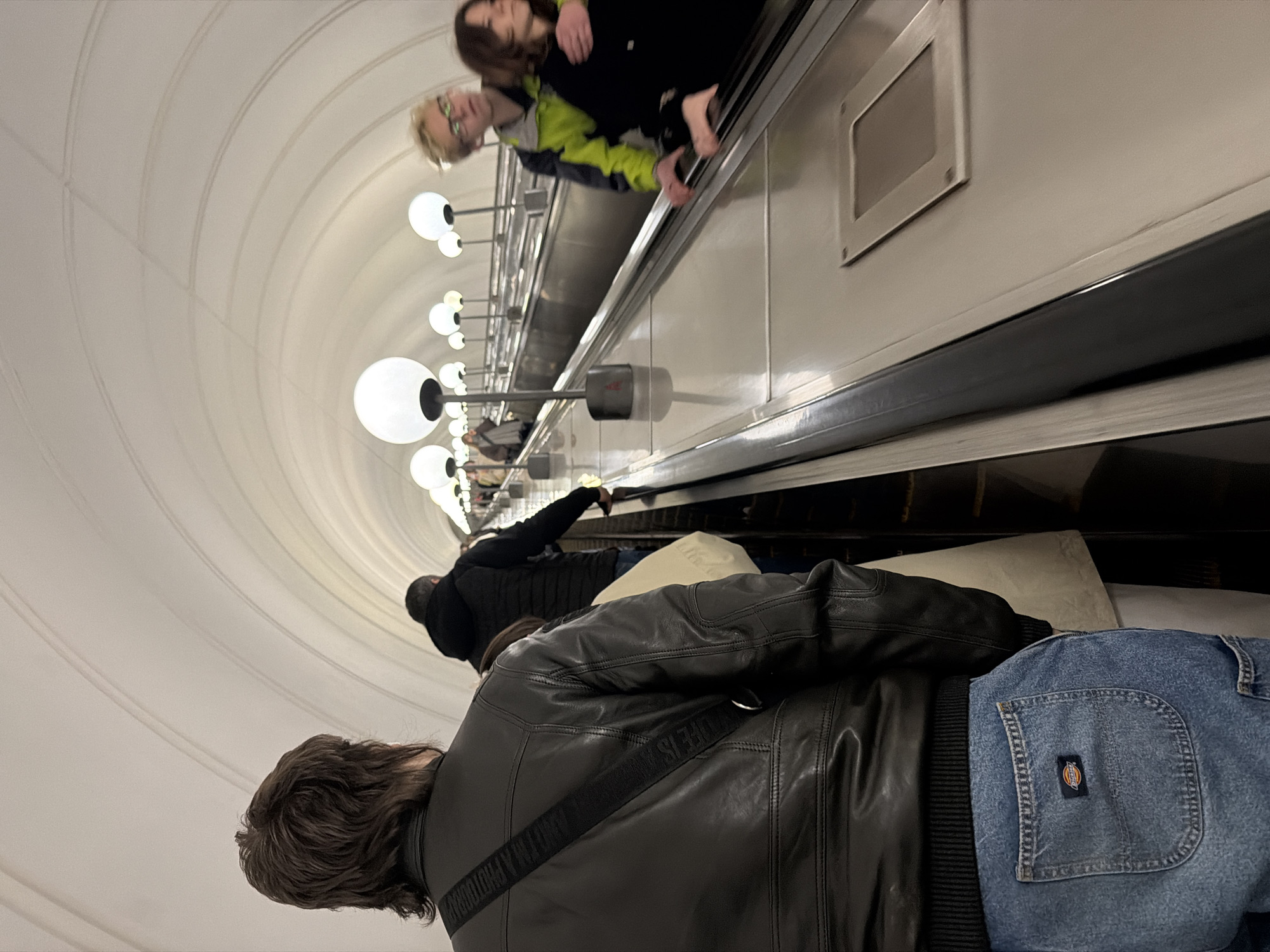 The image depicts an escalator tunnel within a Moscow Metro station, showcasing its unique architecture and people riding the escalator. The perspective is from behind individuals moving up the escalator, looking towards the top.
In the foreground, a person with dark, wavy hair is seen from the back, wearing a black leather jacket with a ribbed waistline. A black strap with white text, partially visible as "I AM A PHOTOGRAPHER," runs across their back. They are wearing light blue denim jeans with a distinct orange and blue "Dickies" brand label clearly visible on the right rear pocket. Further ahead on the escalator, another person is visible from behind, clad in a dark, possibly black, hooded jacket or coat.
At the top of the escalator, two individuals are seen, one with light-colored hair and glasses wearing a lime green and grey jacket, and another with darker hair. Their reflections are also visible on the polished metallic surfaces of the escalator's side panels.
The setting is characterized by a high, curved, and paneled ceiling, painted in a uniform light, off-white color, creating a sense of grandeur typical of Moscow Metro stations. Numerous large, spherical white light fixtures, arranged individually and in pairs, are mounted along the ceiling and side walls, providing bright illumination to the tunnel. The escalator itself features dark handrails and sleek, grey metallic side panels. A rectangular vent or panel is visible on the upper section of the escalator wall. The scene captures the daily commute within an iconic underground transport system.