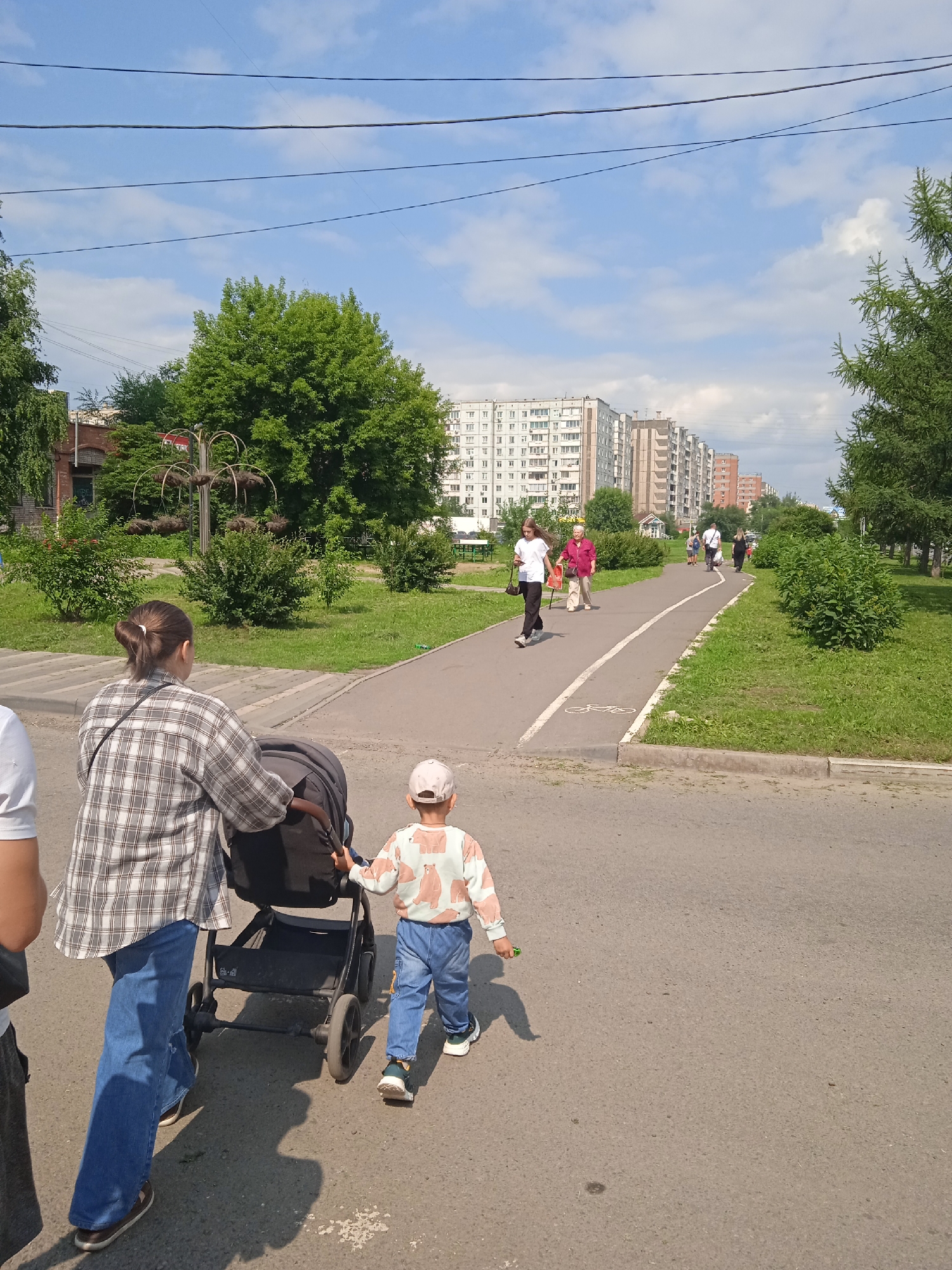 The image depicts a sunny day in a city park with apartment buildings in the background. In the foreground, a woman wearing a plaid shirt and blue jeans pushes a stroller. A young child in a patterned shirt and blue jeans walks beside her. Other pedestrians are visible on a paved path in the middle ground. The park area features green grass, trees, and bushes. The context of Krasnoyarsk, Russia is not directly discernible from the visual elements alone.