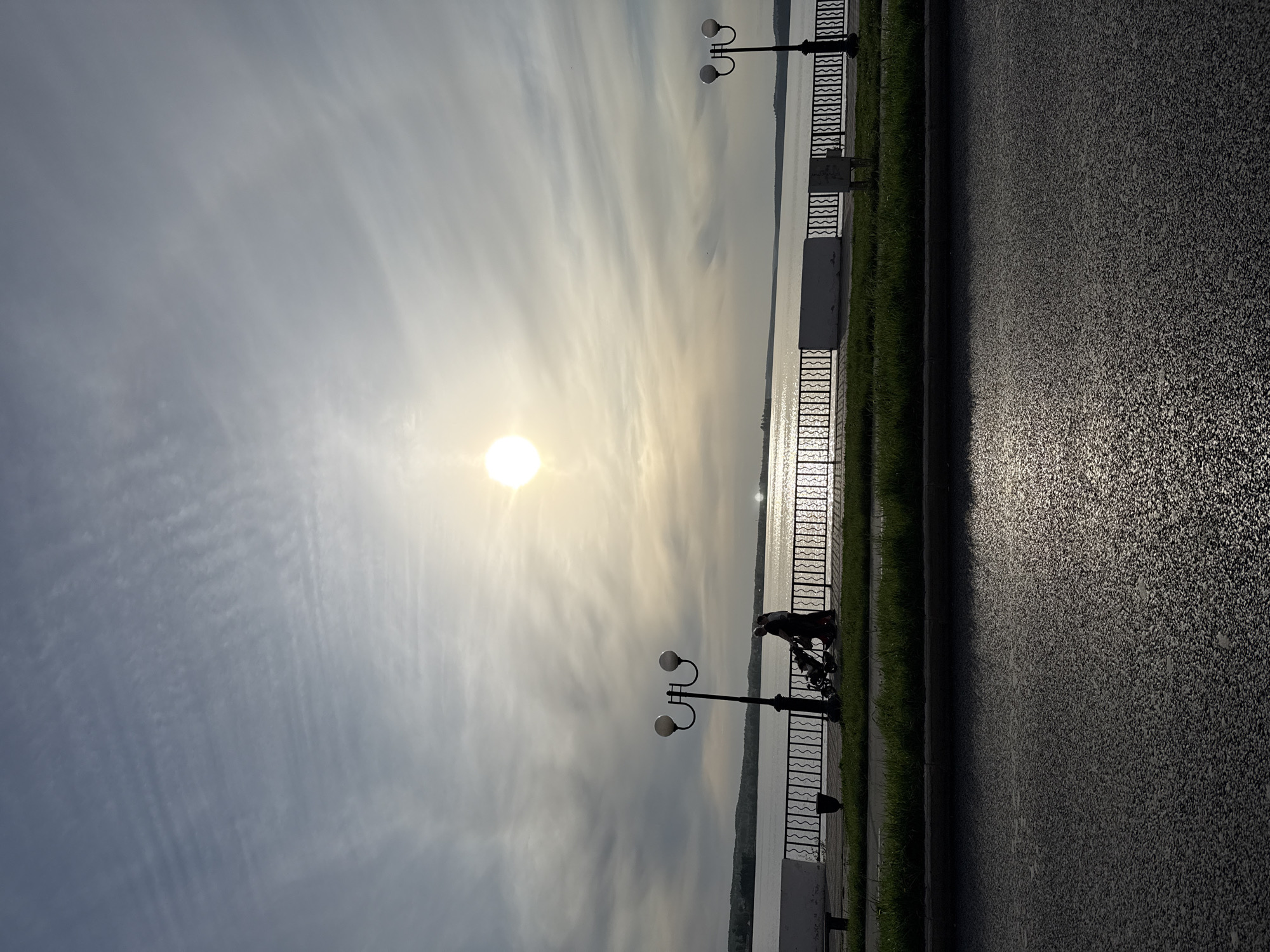 The image shows a scene on the Votkinsk reservoir embankment in the city of Votkinsk, Russia. The sun is prominent in the sky, with bright reflections on the water. A black decorative metal fence runs along the embankment, separating it from the water. Along the fence are several black lampposts with spherical lights. A figure and a stroller can be seen beside the fence, close to one of the lampposts. The embankment surface appears to be made of asphalt. Grass grows between the fence and the embankment surface. The sky is partly cloudy.