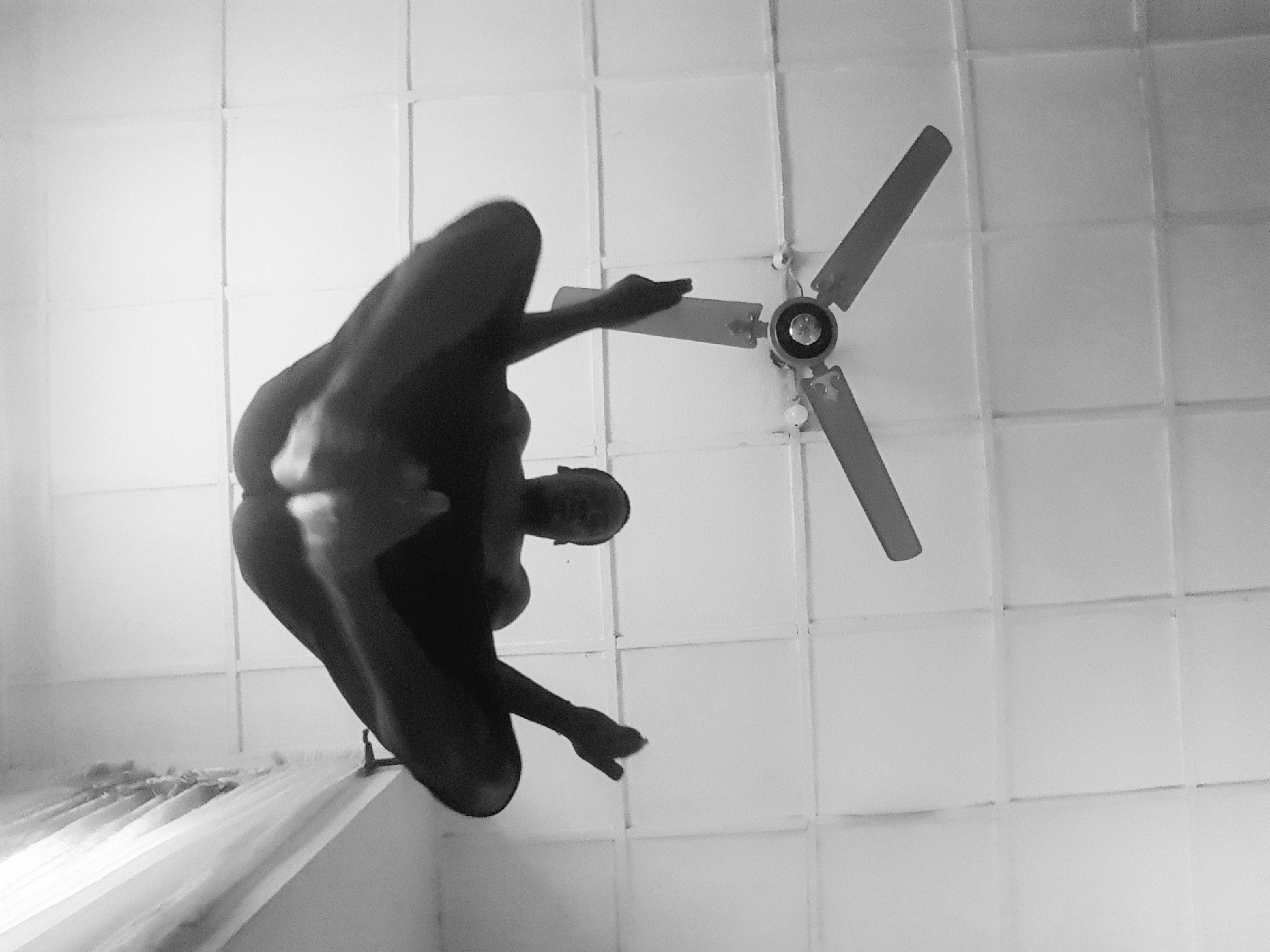 An upward-facing, black and white, wide shot captures a person in mid-air, seemingly suspended from a ceiling fan. The person, whose gender is not discernible, is positioned with their legs bent and spread, and arms outstretched, as if in a yoga pose or diving into an unseen pool. The individual is dark-skinned and appears nude, with their body silhouetted against the bright, tiled ceiling. The ceiling fan, with three blades, is partially visible above the person, with its central motor unit and lights also in view. The room's interior is characterized by a grid-like pattern on the ceiling, suggesting a conventional room. To the right of the frame, a portion of a white curtain or blinds is visible, adding a subtle contrast to the otherwise stark environment. The lighting in the image is diffused, casting soft shadows and highlighting the texture of the ceiling tiles. The perspective suggests the photograph was taken from a low angle, looking directly up at the ceiling and the suspended individual.
