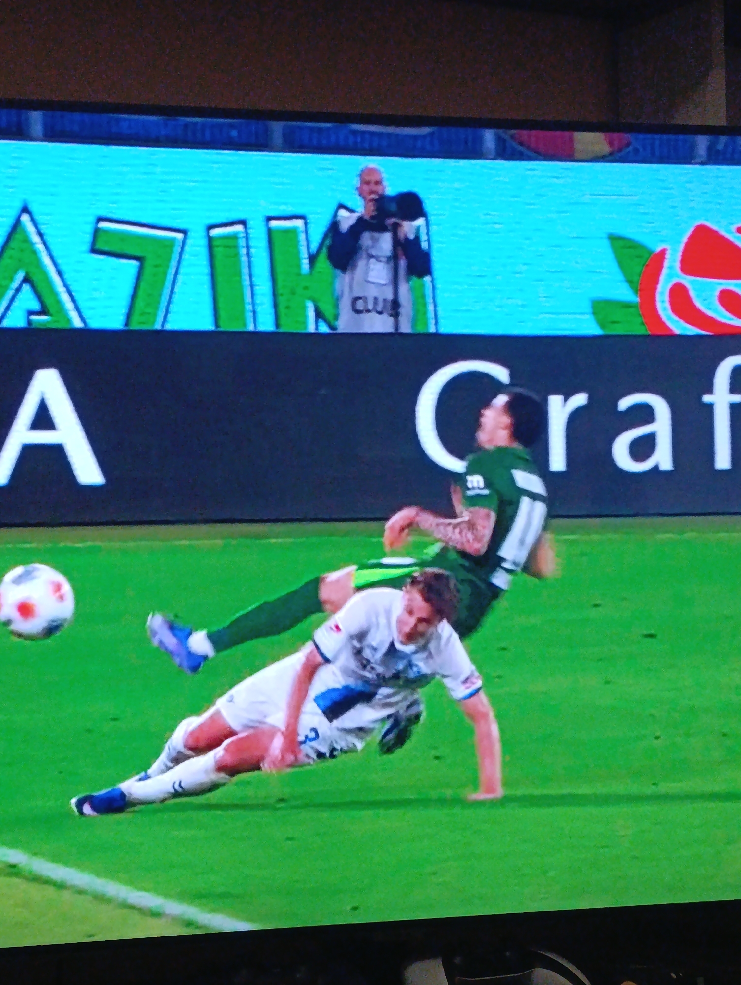 A soccer match in Plovdiv, Bulgaria, showing two players in action on a green field. One player, wearing a white uniform with blue accents, is on the ground. The other player, in a green uniform with white accents, is airborne in what appears to be a kicking motion. A soccer ball is also in mid-air. In the background is stadium advertising with a rose motif and the text "AZIK" and "Craft". A person holding a microphone is visible above the advertising.