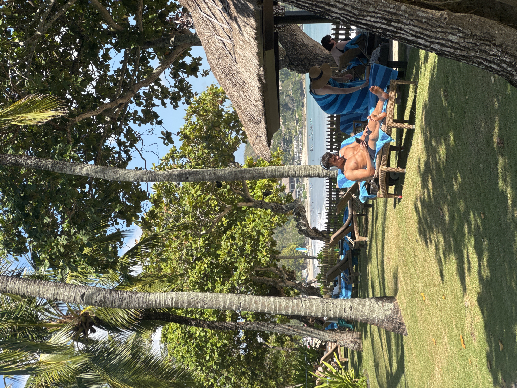 The image depicts a tropical beach scene in Kuta Selatan, Indonesia. In the foreground, a young man with headphones on is lying on a lounge chair, looking at his phone. He is wearing dark swim trunks and has a muscular physique. Adjacent to him, another person wearing a straw hat and a blue and white striped towel is leaning over a lounge chair. Further back, another person is visible on a lounge chair. The lounge chairs are situated on a manicured green lawn with scattered fallen leaves. Towering palm trees and leafy green trees provide shade. In the background, a body of water, likely the ocean, is visible with several small boats or surfboards on it. The shoreline is lined with buildings and vegetation. The sky is clear and blue. The overall atmosphere is relaxed and leisurely, characteristic of a beach resort.
