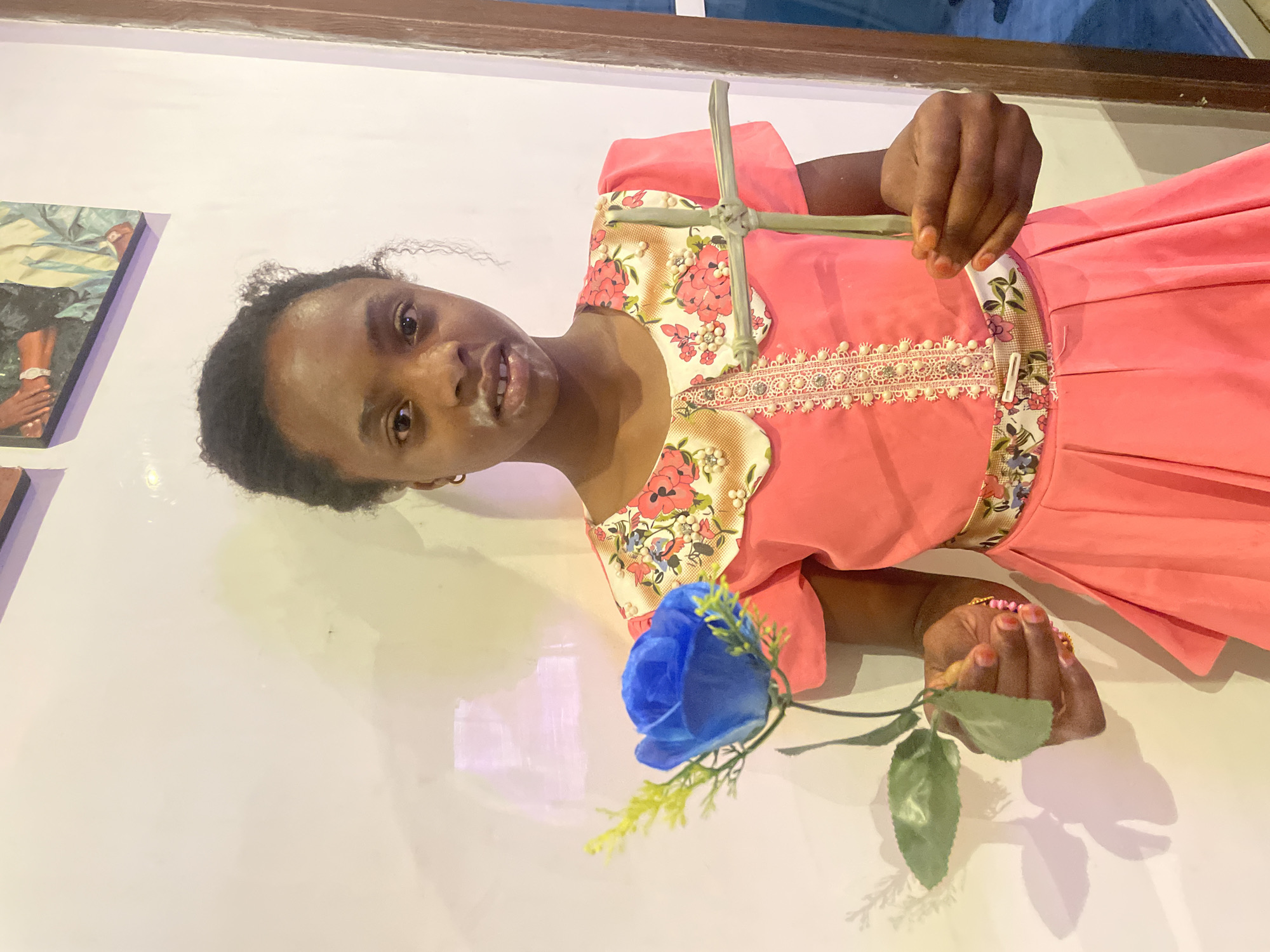 A woman with dark hair and skin is holding a blue rose in her right hand and a palm leaf cross in her left hand. She is wearing a pink dress with floral trim. The location is Jos, Nigeria. The background is a white wall. There is a painting on the wall to the left of the woman.