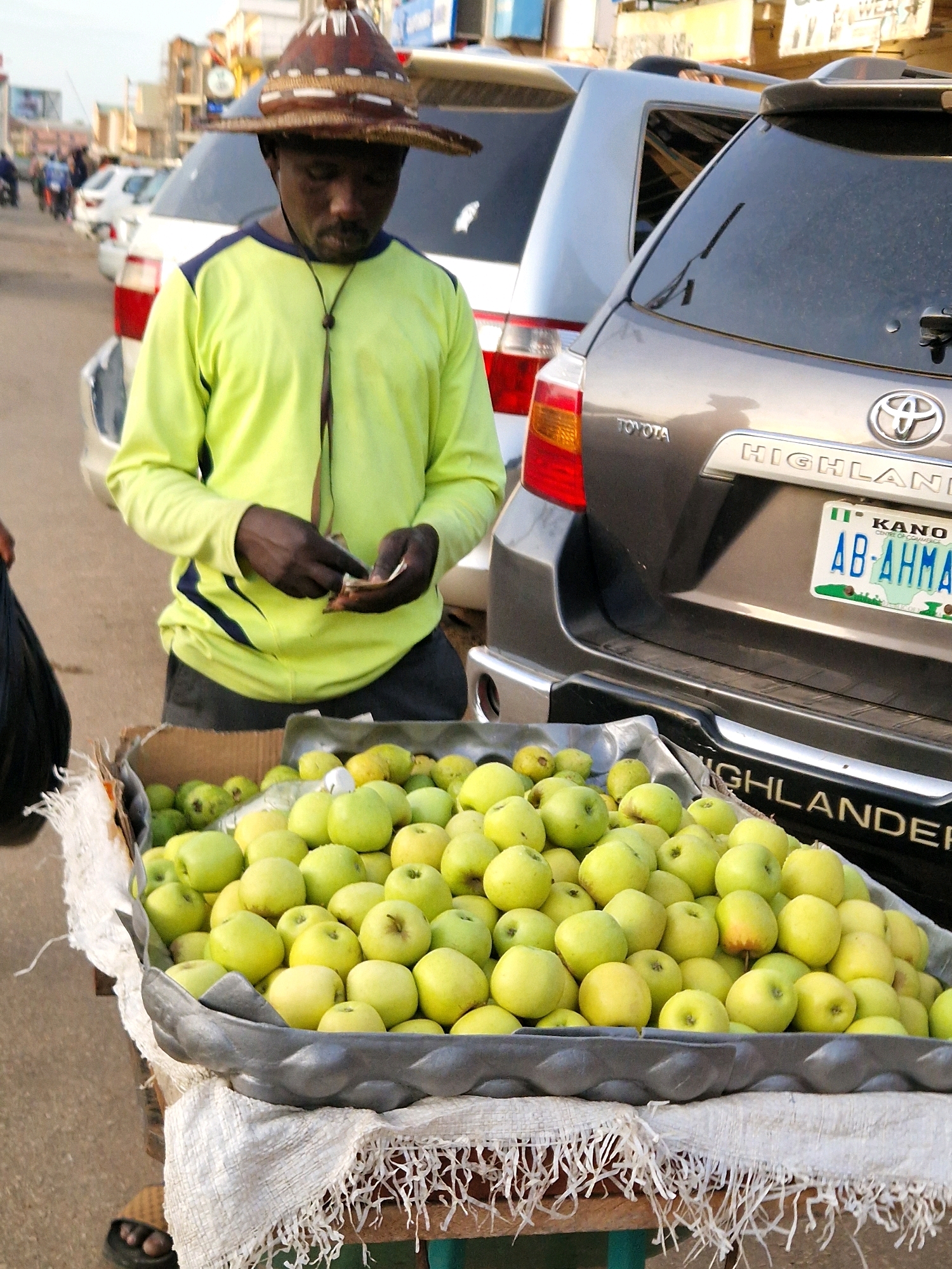 In Jos, Nigeria, a street vendor is seen standing next to a cart filled with green apples. He wears a yellow long-sleeved shirt and a traditional patterned hat. His hands are engaged in counting money. Behind him, a silver Toyota Highlander SUV is parked, displaying a Nigerian license plate from Kano. The scene depicts a typical street market environment with other vehicles and buildings visible in the background, suggesting a bustling urban setting.