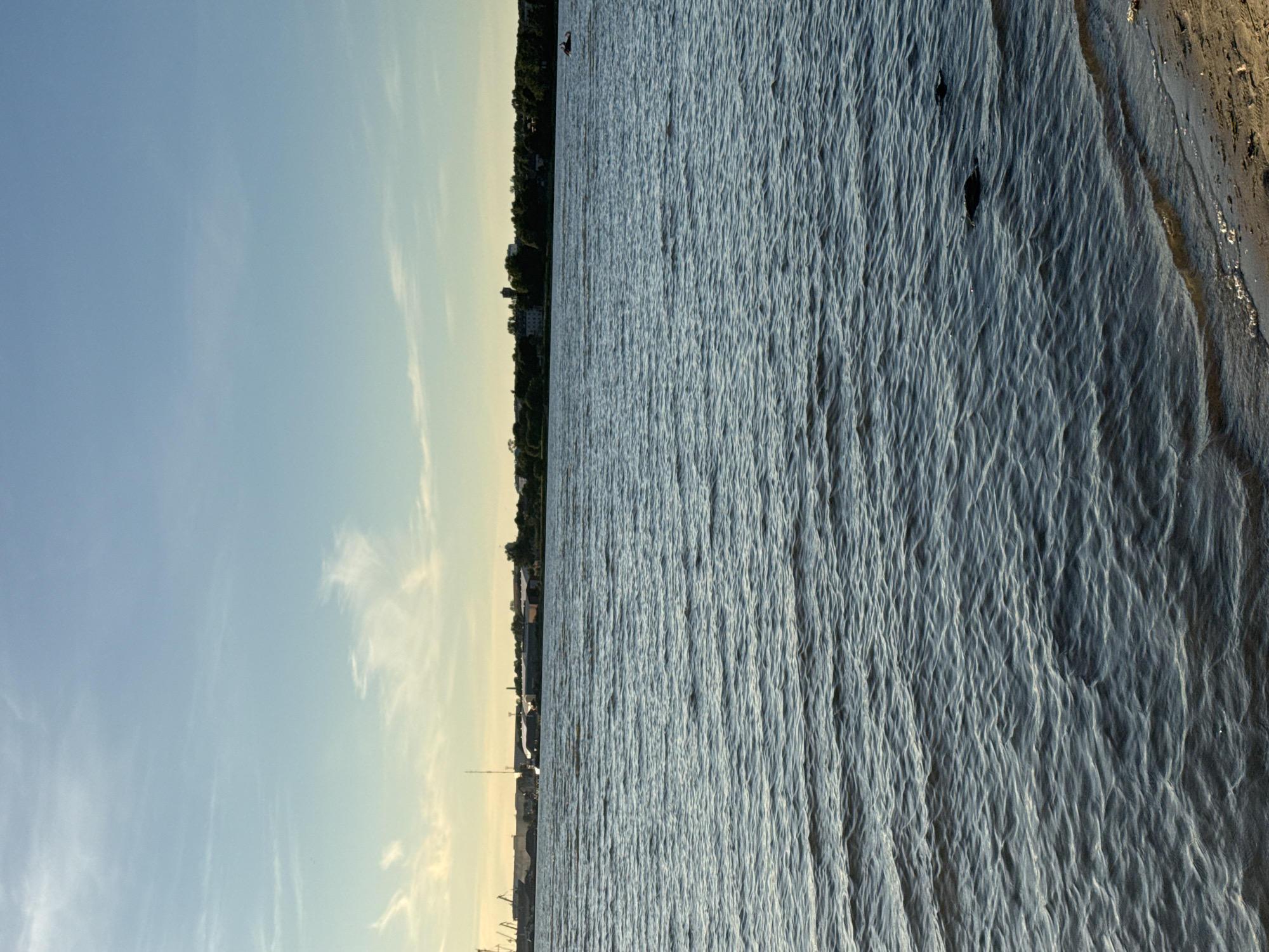 The image depicts a wide body of water with a gentle ripple on its surface, suggesting a calm day. The water occupies the majority of the frame, with a sandy shoreline visible in the lower right corner. In the distance, a cityscape with industrial structures, buildings, and trees lines the horizon under a vast, pale blue sky with wispy clouds. The sky is brighter towards the horizon, indicating either sunrise or sunset. A single jet ski can be seen in the middle distance, leaving a wake on the water. The scene appears to be a view from the shore of the Tallinn Bay, possibly near the industrial port area.