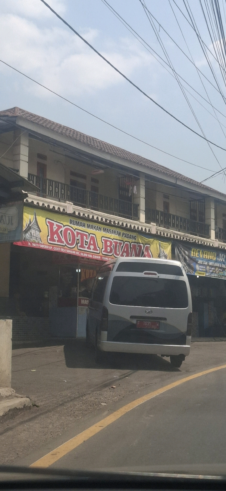 A silver minivan with license plate "F 7035 W" is parked on a street in front of a building with a banner that reads "RUMAH MAKAN MASAKAN PADANG KOTA BUANA". The banner also displays an address: "JL. RAYA CIBEBER KP. CIJATI DESA SUKASARI KEC. CILAKU KAB. CIANJUR". The building appears to be a two-story structure with balconies. Overhead power lines crisscross the sky, which is partly cloudy. The street in front of the building is paved and has a yellow dividing line. A concrete pillar is visible on the left side of the image.