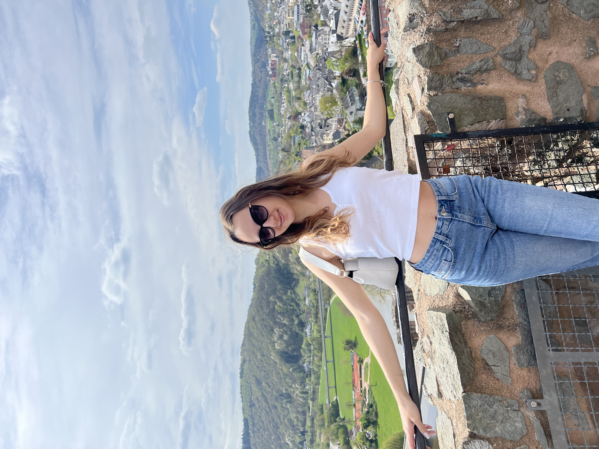 A full-length image shows a young woman standing on a stone structure overlooking Saarburg, Germany. She is leaning against a metal railing, with her left arm extended towards the foreground and her right arm behind her. She wears sunglasses, a white tank top, and blue jeans. A white shoulder bag is visible. The background features a view of Saarburg, including the city's buildings, a wooded hill, and the Sarre River, under a sky with scattered clouds. The stone structure on which she stands has a metal grate attached to it.