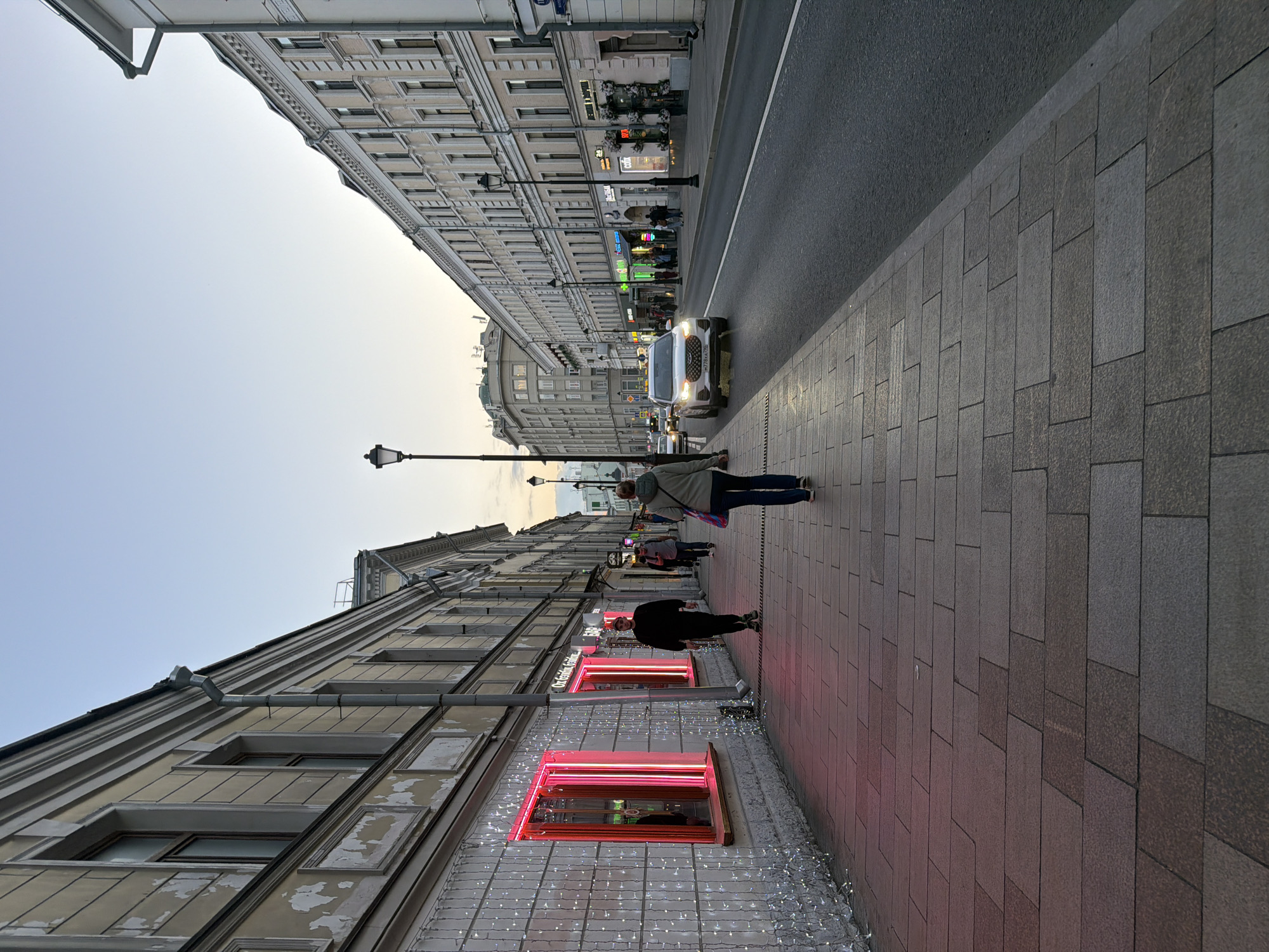 The image depicts a street scene in Moscow, Russia. The buildings are tall and appear to be made of stone or concrete, with a light beige or gray color. The street is divided into a road for cars and a sidewalk for pedestrians. There is a street lamp visible on the side of the road. There are pedestrians walking along the sidewalk. A light-colored car is traveling on the road. On the bottom left, building is decorated with lights and has windows with pink neon outlines. The sky is light in color, suggesting it may be daytime or dusk.