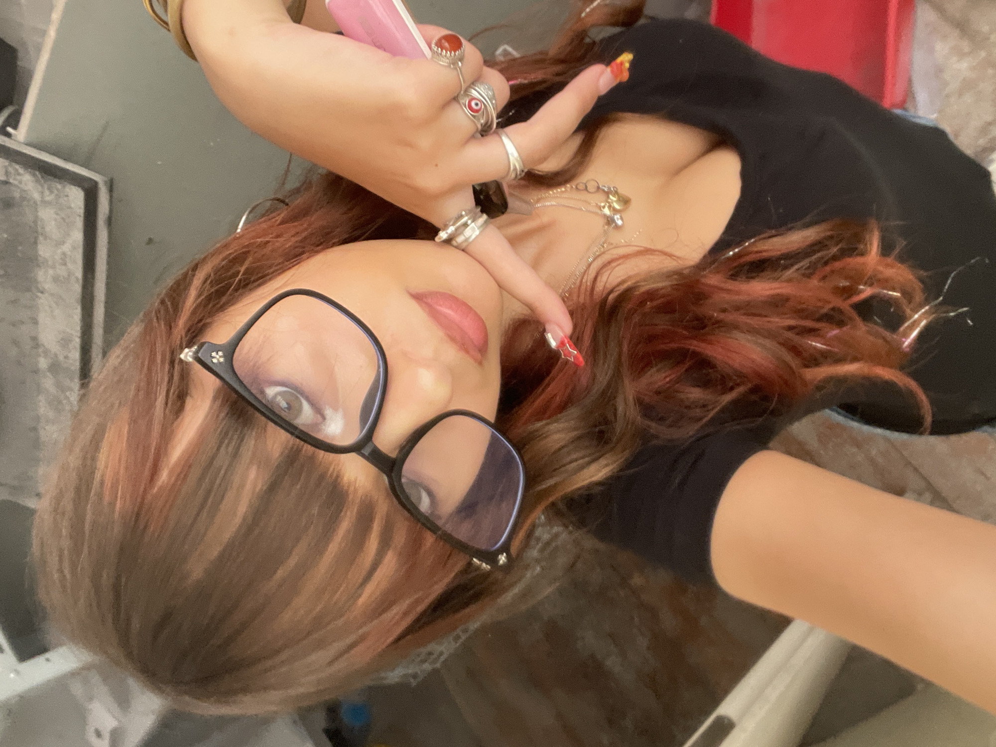 A young woman with long, auburn hair and bangs, wearing black-rimmed glasses, a black t-shirt, and multiple silver rings on her fingers, poses for a selfie. Her right hand is raised, with two fingers extended, holding a pink object. She is also wearing several necklaces, including one with a heart-shaped pendant. The background appears to be an indoor environment with some shelving units and a floor that seems to be made of concrete or wood. The image does not contain any discernible landmarks or features that would indicate a specific location within Raanana, Israel.