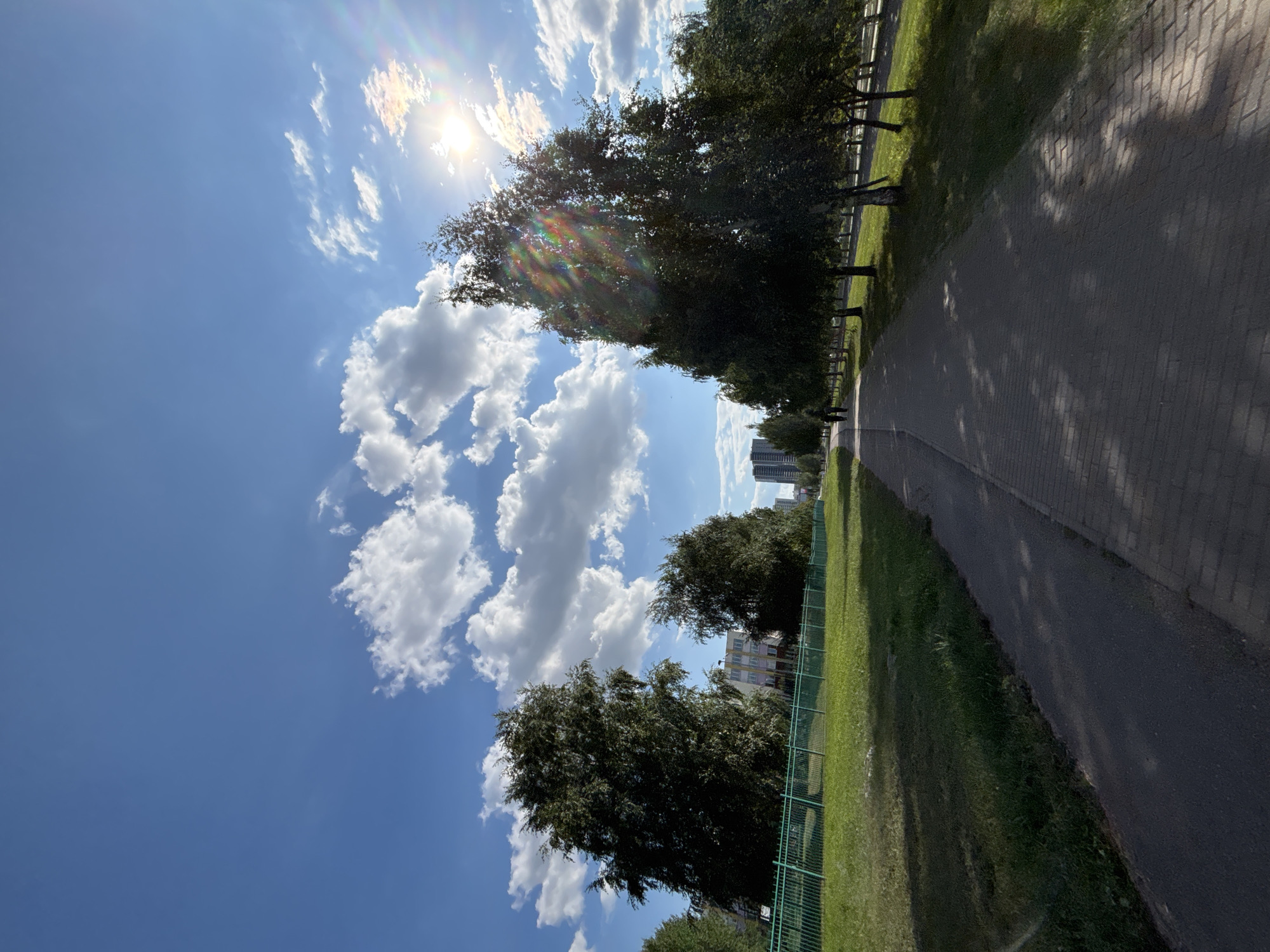 A sun-drenched paved pathway, lined by trees and grass, runs through an urban park in Naberezhnye Chelny, Russia. The pathway is made of interlocking rectangular bricks and is partially shaded by the leaves of the trees. The grass is green and well-maintained, with a green metal fence bordering it on the left side of the image. In the distance, several apartment buildings and a tall skyscraper are visible, indicating the park is located within a residential area. The sky is a vibrant blue with scattered white, fluffy clouds. The sun is shining brightly, creating a lens flare effect in the upper right corner of the image. Two figures can be seen walking away from the camera on the pathway in the distance.