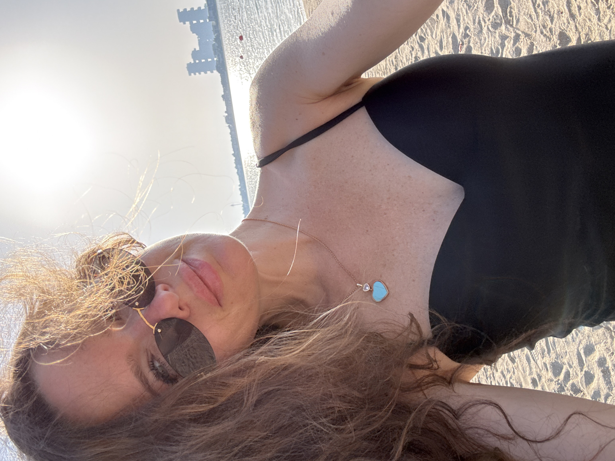 A woman with long, wavy brown hair, wearing sunglasses and a black swimsuit, takes a selfie on a sandy beach. A turquoise heart pendant necklace adorns her neck. In the background, the sun glints off the water, and distant buildings rise from the Dubai skyline. The scene appears to be a bright, sunny day.