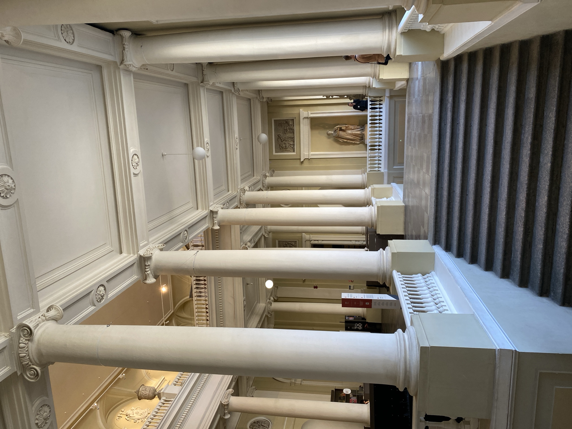 The image depicts a long hallway lined with classical columns, likely an interior space within a museum or government building. The columns are white and appear to be of the Doric or Ionic order, featuring decorative capitals. On the left side, ornate white walls with detailed moldings and decorative medallions are visible. On the right, a grey stone staircase rises. At the end of the hallway, artwork and sculptures are displayed on the walls. Several people are present, standing and walking along the hallway. Artificial light sources, such as hanging lamps, illuminate the space.