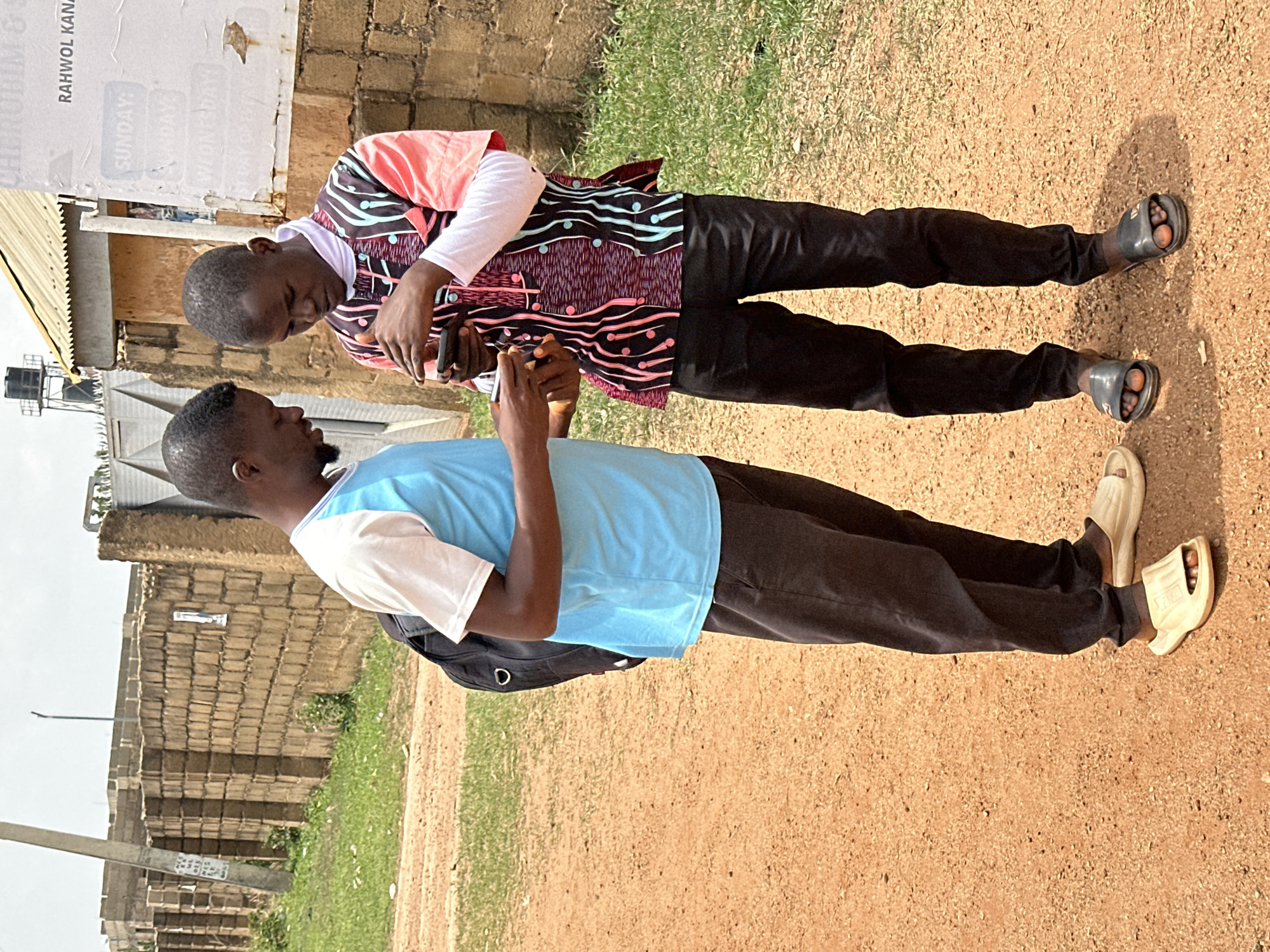 Two men are standing outdoors in Bukuru, Nigeria, both holding mobile phones and looking at them. One man wears a light blue t-shirt with white sleeves, black trousers, and tan sandals; he has a black backpack on. The other man is wearing a long-sleeved patterned shirt in shades of pink, maroon, and white over a white undershirt, black trousers, and dark grey sandals. They stand on a reddish-brown dirt ground with some green grass growing in patches. A brick wall is in the background to one side, and a building with a sign on it is visible in the background on the other side. The lighting suggests it is daytime.