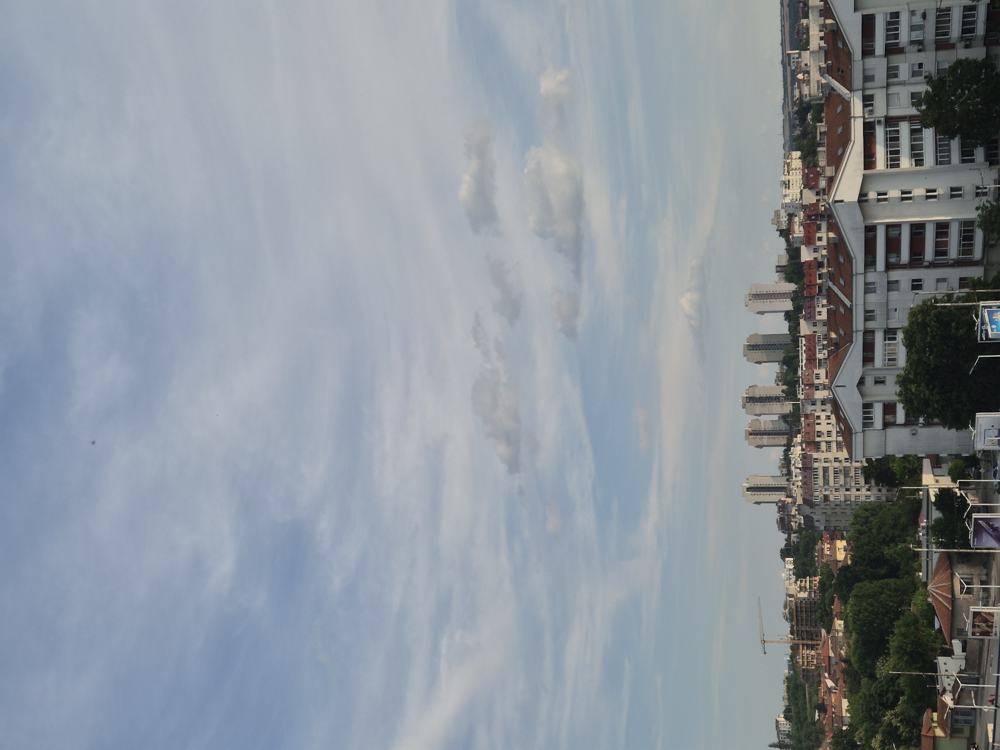 The image shows a panoramic view of a cityscape under a partly cloudy sky. In the foreground, residential buildings with tiled roofs and white facades are visible. The middle ground features a mix of buildings, including apartment complexes and smaller houses, interspersed with green trees. In the background, several tall, modern apartment buildings stand out against the horizon, along with a construction crane. The sky is a soft blue with streaks of white clouds, suggesting a mild day. The lighting indicates it could be late afternoon or early morning. The scene is devoid of any prominent human presence or activity. The setting appears to be an urban or suburban environment.