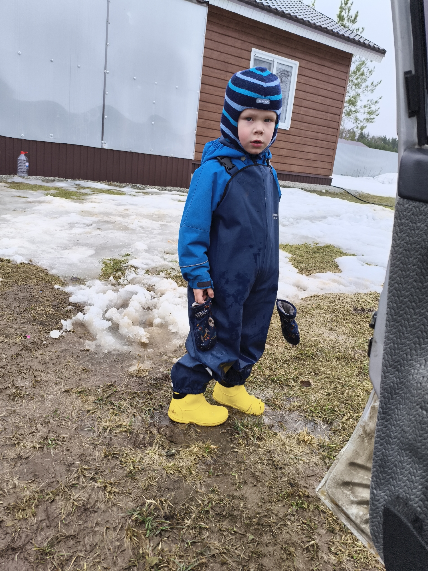 A young child wearing a blue jumpsuit, blue jacket, blue and white striped hat, and yellow boots stands in a muddy yard. The child is holding two dark colored mittens. The yard has patches of grass and melting snow. Behind the child is a building with white siding and brown trim. To the right of the child is a grey vehicle. The image was taken in Русский Вожой, Russia.