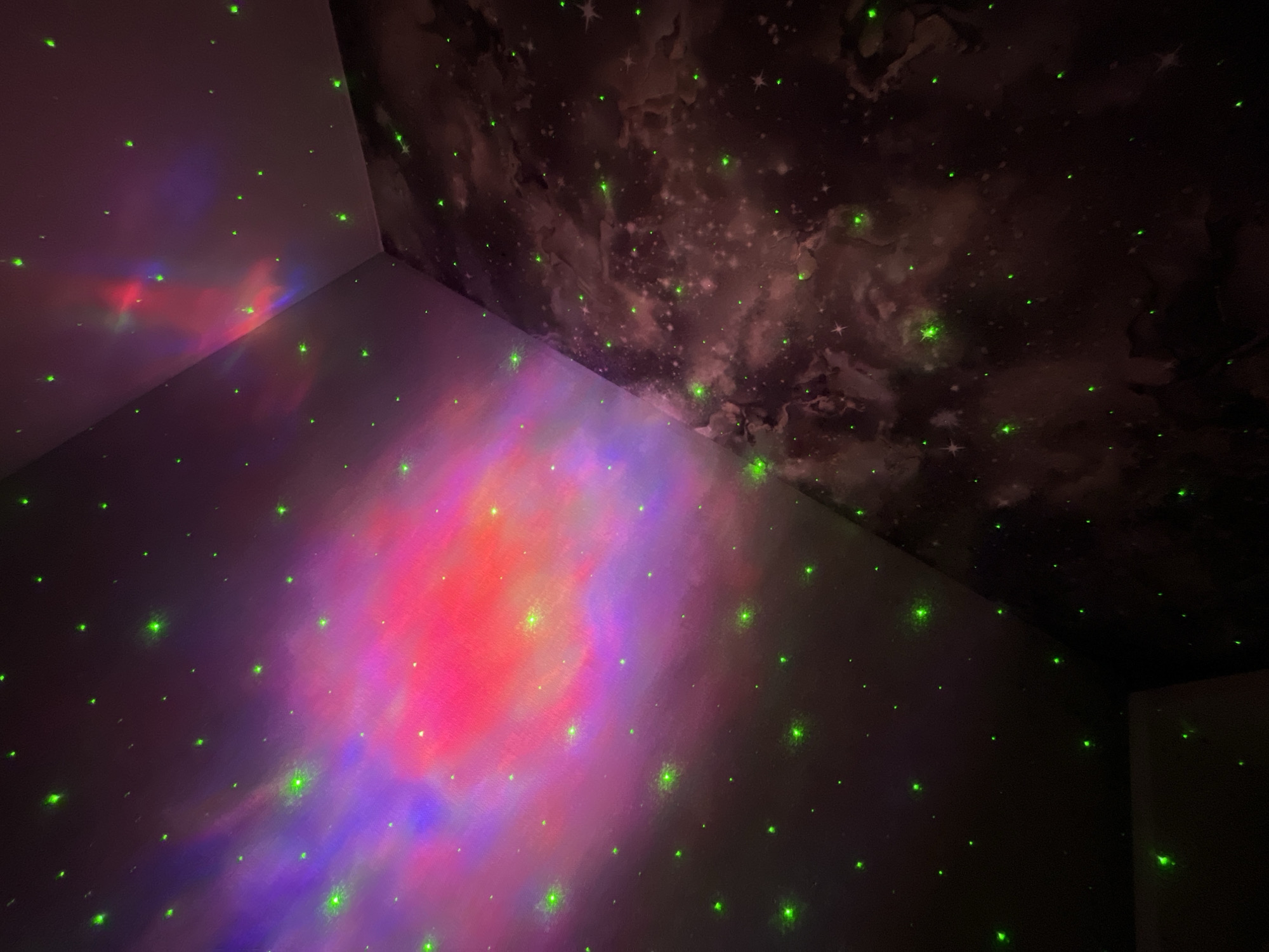 This is a photograph of a room with a projector casting a nebula and star effect onto the walls and ceiling. The nebula is predominantly pink and purple, with smaller green lights scattered across the surfaces, simulating stars. The room itself appears to be a corner, with one wall featuring a darker, cloud-like pattern. There are no people or discernible objects other than the projector's light display. The location is not specified within the image or its context.