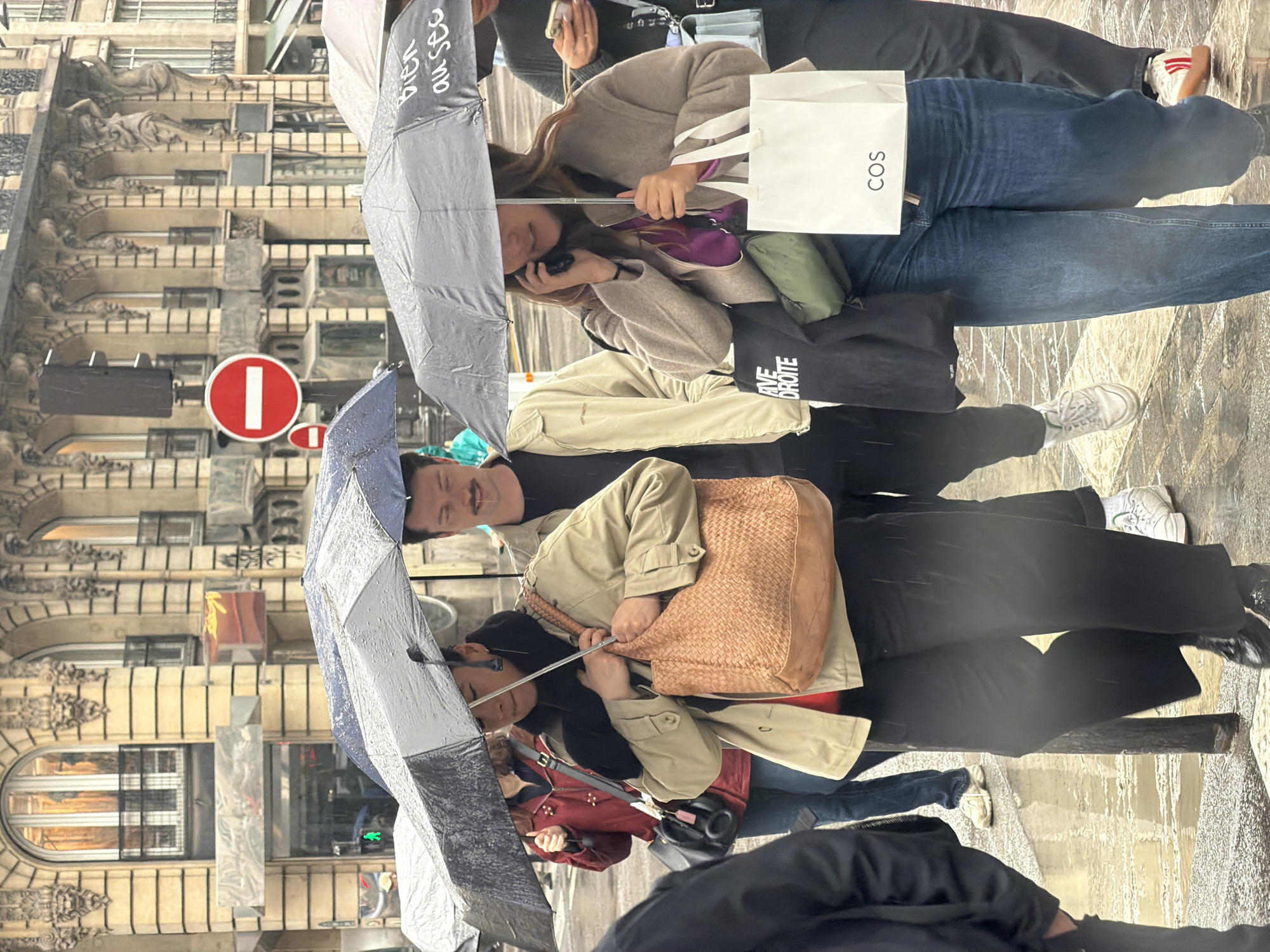 On a wet street in Paris, a group of people are walking under umbrellas in the rain. The architectural style of the buildings suggests the location is Paris, France. Several people are huddled under umbrellas, and at least one is carrying a shopping bag from "COS". Street signs are visible in the background. One person is looking up, smiling.