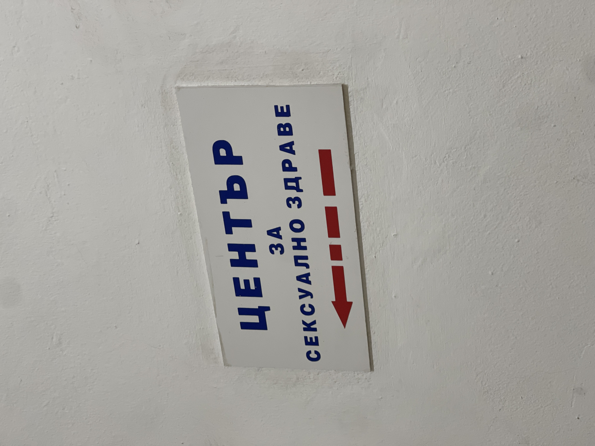 The image shows a sign on a white wall, written in Bulgarian, indicating the direction to the "Center for Sexual Health" in Plovdiv, Bulgaria. The sign is white with blue lettering and a red arrow pointing to the right.