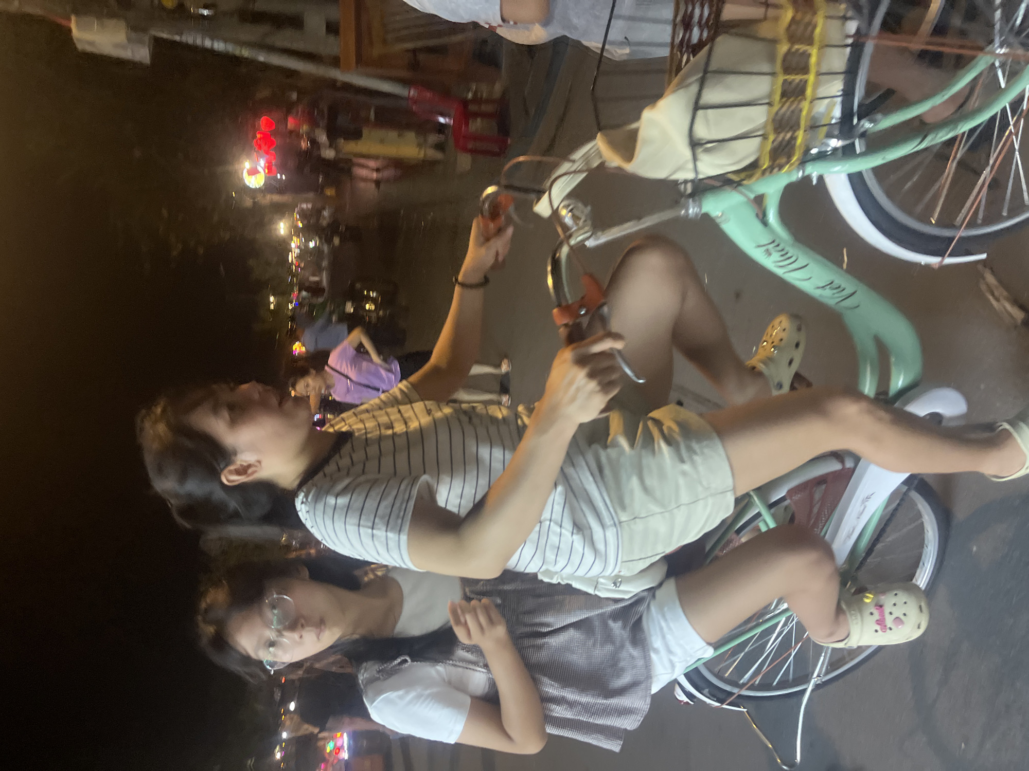 In Hội An Ward, Vietnam, at night, two young women ride a bicycle side-by-side. The woman in front, wearing a striped t-shirt and beige shorts, pedals the bike. The woman behind her, wearing glasses, a white t-shirt, and a patterned vest over white shorts, holds onto the back of the seat. They are on a street with other people and vehicles visible in the background, illuminated by streetlights and glowing red lanterns. The bicycle is light green and has a basket on the front.