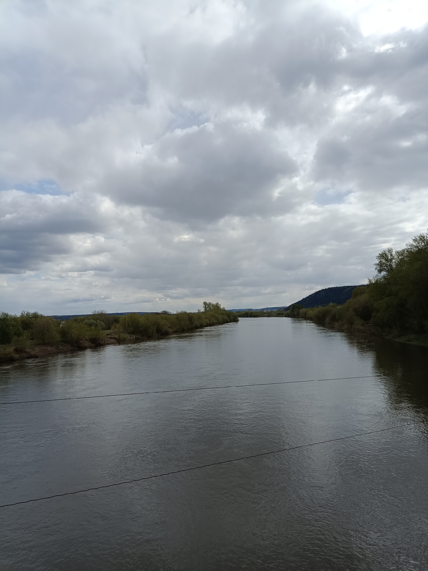 The image depicts a wide river flowing through a landscape under a cloudy sky. The river is dark and reflects the light of the sky. On either side of the riverbank, there is a line of trees and vegetation. In the distance, a small hill rises above the trees on the right side of the river. Overhead is a sky filled with layers of gray clouds. There are power lines stretched horizontally across the lower portion of the image. This scene is located in Посад, Russia.
