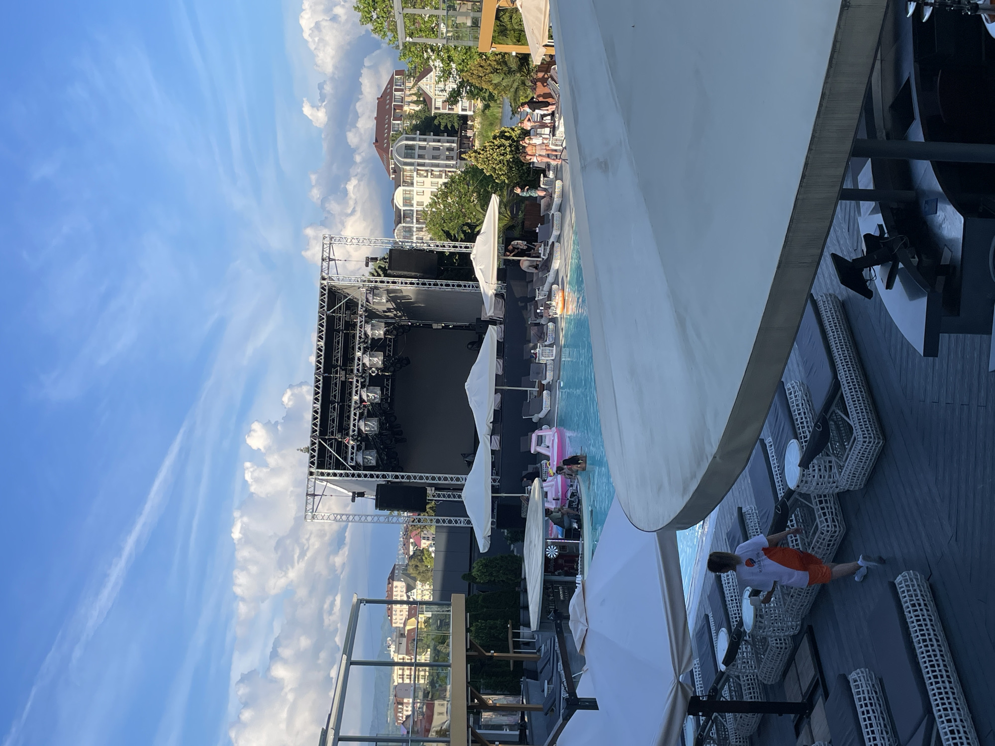 The image depicts an outdoor venue with a swimming pool and a stage set up in Sochi, Russia. The scene is viewed from a slightly elevated position, looking towards the stage and the pool area.
In the foreground, a large white umbrella partially obscures the view. Beyond this, a dark wooden deck is covered with rows of white wicker lounge chairs with dark cushions. Some of these chairs are occupied. A person wearing an orange shirt and shorts is walking away from the camera across the deck, carrying a white cup.
The swimming pool occupies a central part of the scene. It is surrounded by more lounge chairs and white umbrellas. Several people are in the pool, including some on inflatable toys. A pink inflatable flamingo is visible. A swim-up bar with a circular counter is situated in the pool, with a few people seated at it.
A large stage with lighting rigs and speakers is positioned at the far end of the pool area. Behind the stage, there are several multi-story buildings with light-colored facades, suggesting an urban or resort setting. Trees and greenery are visible between the buildings. The sky above is blue with scattered white clouds, some of which are large and puffy. The overall impression is that of a daytime scene at a lively outdoor entertainment venue.