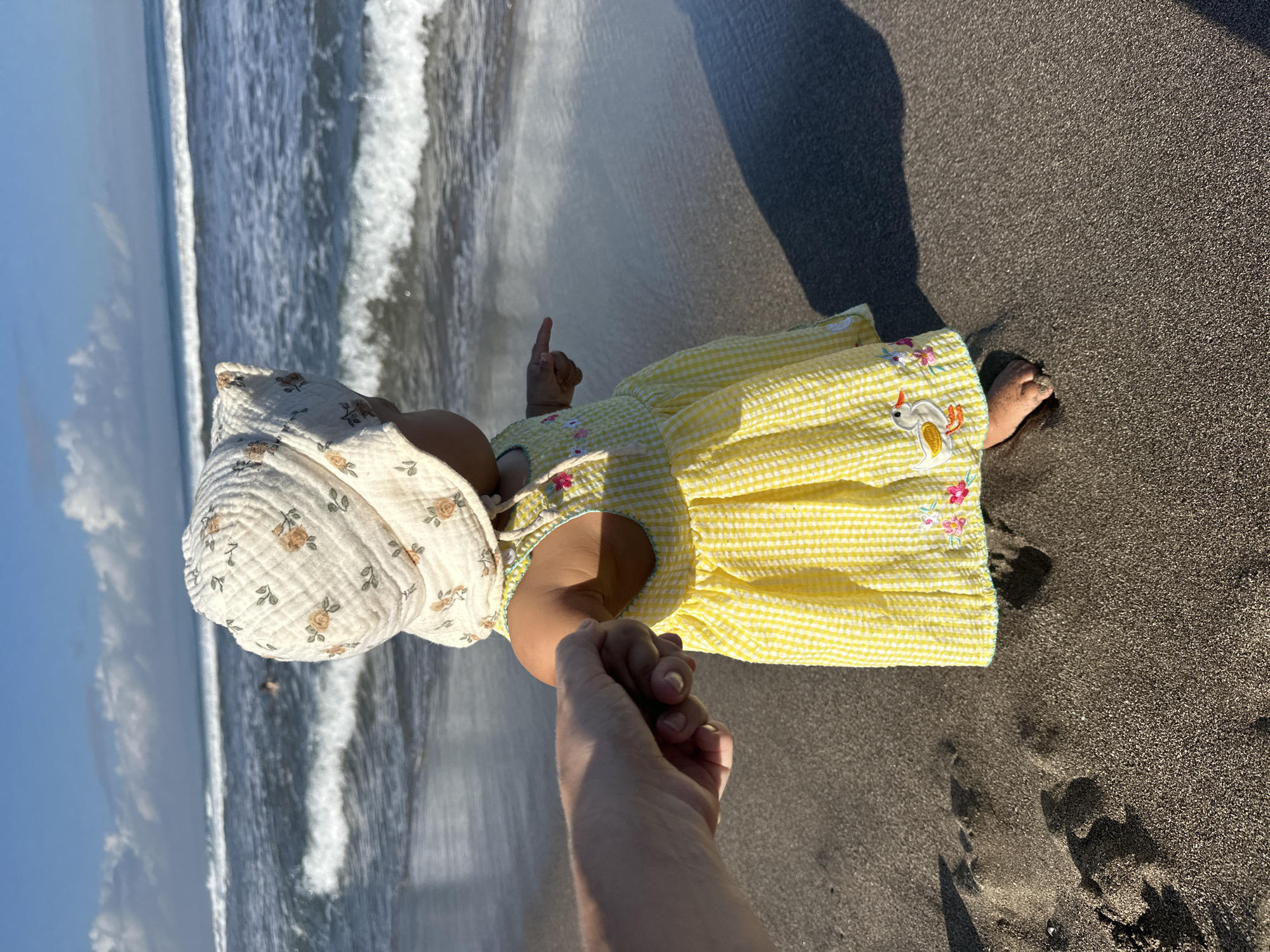 A baby wearing a floral patterned hat and a yellow and white checkered dress with floral and duck embroidery stands on a dark gray sand beach, holding the hand of an adult. The baby points with their other hand toward the ocean, where waves are visible. The scene is brightly lit, casting shadows from both the baby and the adult onto the sand. The location is North Kuta, Indonesia.