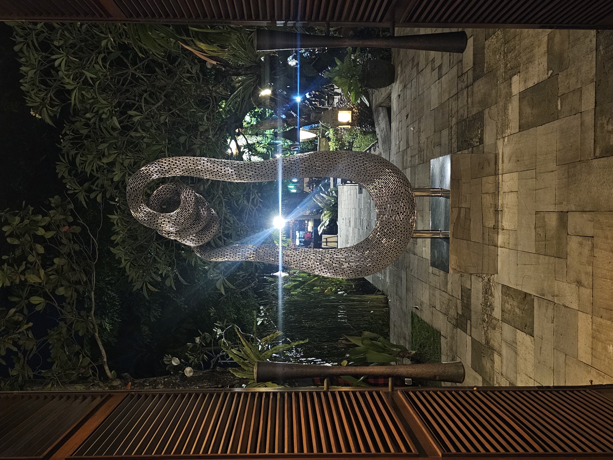The image depicts an outdoor sculpture resembling a woven metal snake in Denpasar, Indonesia. The sculpture is illuminated and positioned above a stone-tiled pathway amidst lush tropical vegetation. The perspective is from below, looking up at the sculpture through what appears to be a wooden slat structure, possibly a pergola or awning. A variety of light sources, including streetlights or decorative lighting, can be seen in the background. Some blurred human figures are in the mid-ground, possibly pedestrians. The overall scene suggests an urban park or outdoor dining area at night.