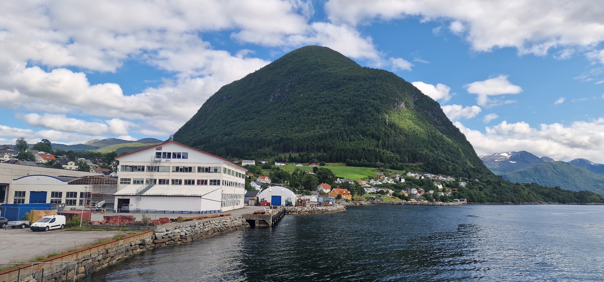 The image captures a waterfront industrial area in Volda, Norway, with a prominent, steep, tree-covered mountain dominating the background. In the foreground, a calm, dark blue body of water laps against a stone embankment and a pier. Several industrial buildings with white siding and red roofs are visible along the waterfront. One large, multi-story building has numerous windows. In the lot next to the buildings, a white van, a trailer, and some large pipes are present. A smaller, rounded blue-roofed building is situated on a pier extending into the water, with a red forklift nearby. Further back, a cluster of residential houses with various colored roofs are scattered on the lower slopes of the mountain. The sky is a bright blue with scattered white clouds. The scene appears to be a working port or industrial zone on the edge of a residential community, nestled against a dramatic natural landscape.