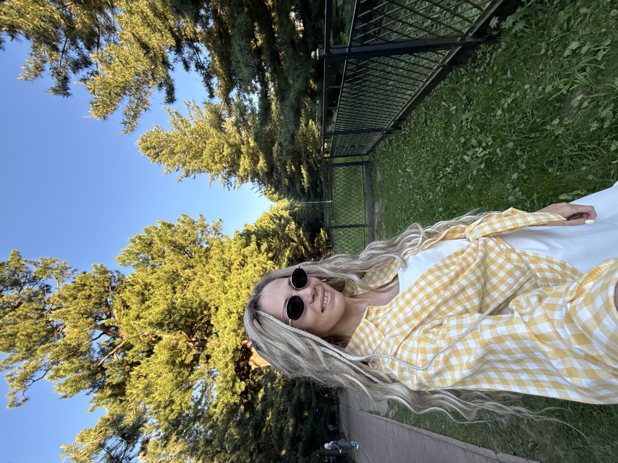 A fair-skinned woman stands in an outdoor park in Yaroslavl, Russia. She has long, wavy blonde hair with several thin white braids incorporated into the style, and wears dark oval sunglasses with thin silver frames. Her attire includes a white top or dress, and a yellow and white gingham-patterned long-sleeved shirt tied at the waist. Her fingernails are manicured and painted white.
The woman is positioned slightly left of center, looking towards the viewer with a slight smile. To her left, a paved pathway recedes into the background, where two blurred individuals are visible: one person in a headscarf facing away, and another figure further down the path.
The background features numerous tall coniferous trees with green foliage, silhouetted against a clear blue sky. A black metal fence, constructed with a diamond lattice pattern and topped with decorative finials, extends along the right side of the frame, dividing a grassy area from the background trees. The ground in the foreground is covered with green grass and some visible weeds. The scene is illuminated by bright natural light, indicating daytime conditions.