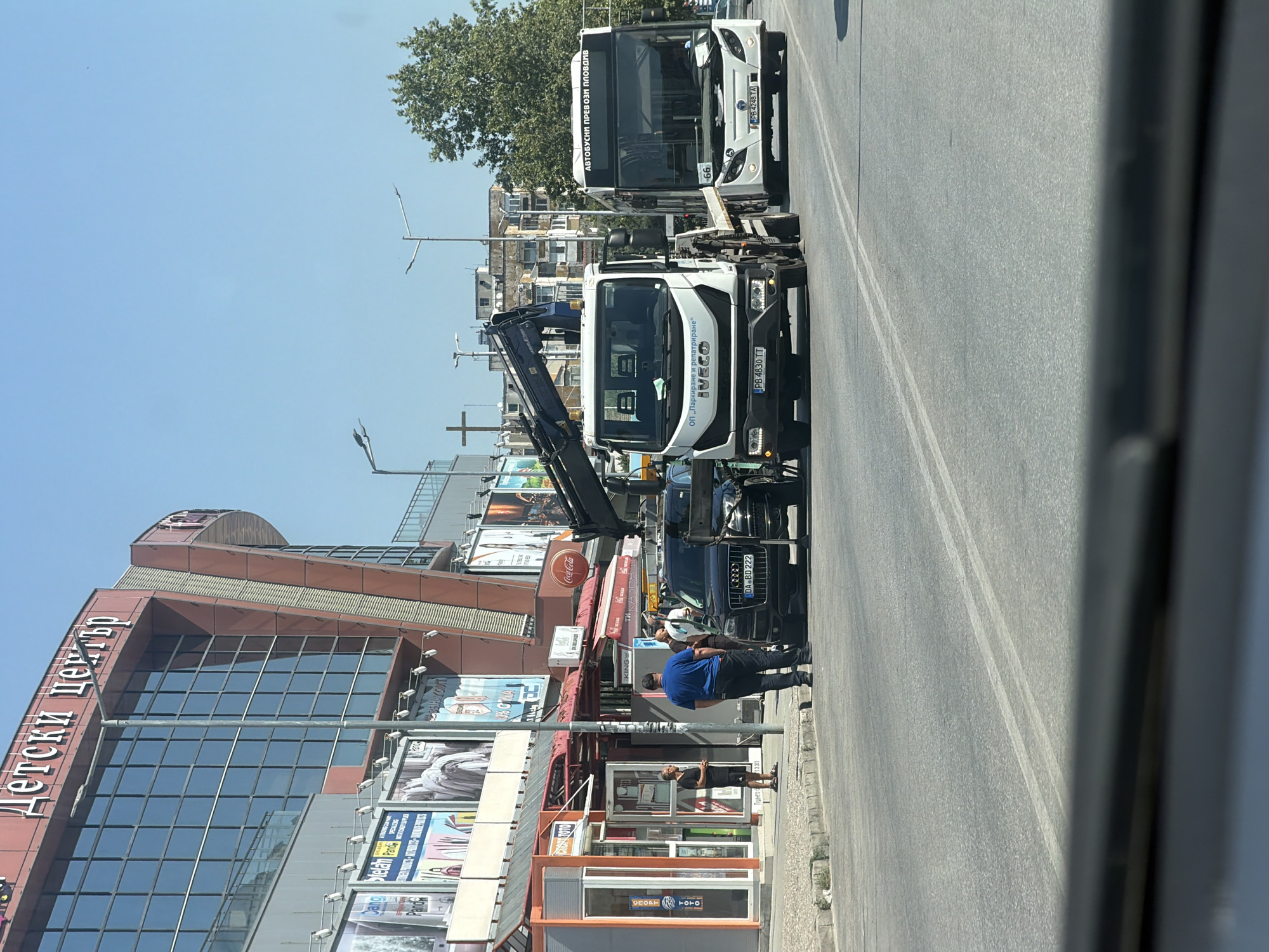 The image depicts a street scene in Plovdiv, Bulgaria. In the foreground, a dark-colored Audi with license plate AB BD 222 is parked on the side of the road, with its front end partially lifted by the crane of a white Iveco tow truck. The tow truck, labeled "ОП „Паркиране и репатриране“ IVECO", is positioned behind the Audi. A white bus with the inscription "АВТОБУСНИ ПРЕВОЗИ ПЛОВДИВ" and license plate PB 4248 TA is visible on the right side of the road, driving past the scene. Several individuals are standing on the sidewalk next to the Audi and the tow truck, appearing to observe or be involved in the situation. Behind them, a building with large windows and signage that reads "ДЕТСКИ ЦЕНТЪР" (Children's Center) is prominent. The environment is an urban setting under a clear blue sky, with other buildings and trees visible in the background.