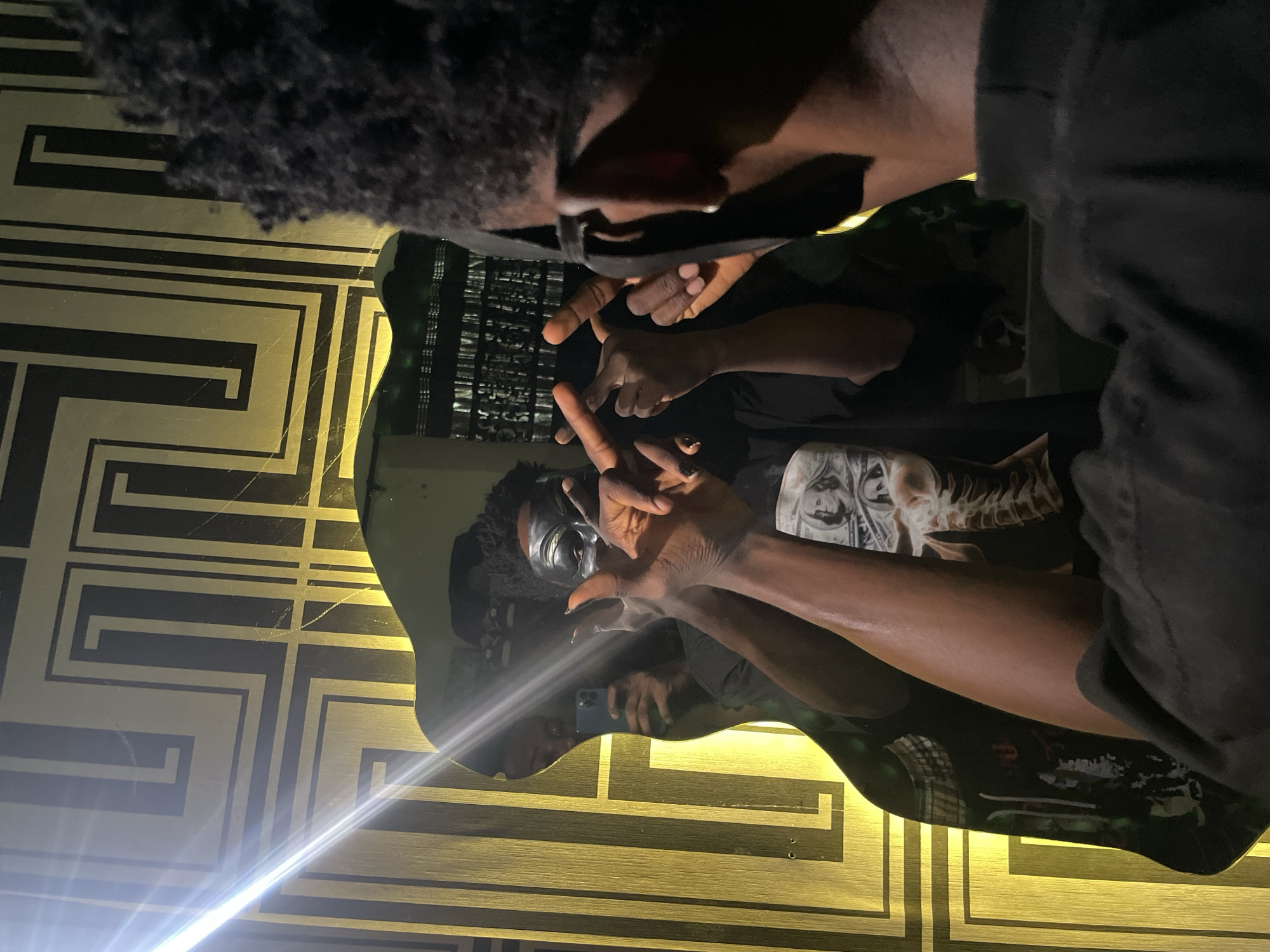The image shows two individuals reflected in a table with a gold and black geometric design. One individual is visible at the top of the frame with dark hair and a black mask pulled down below the nose. Their hands are raised, making a gesture. The second individual is reflected in the table, wearing a dark shirt with a graphic design and dark headwear. Their hands are also raised in a similar gesture. A phone is visible in the reflection, held by an unseen person. The location is not definitively identifiable as Lokoja, Nigeria, based solely on the visual information.