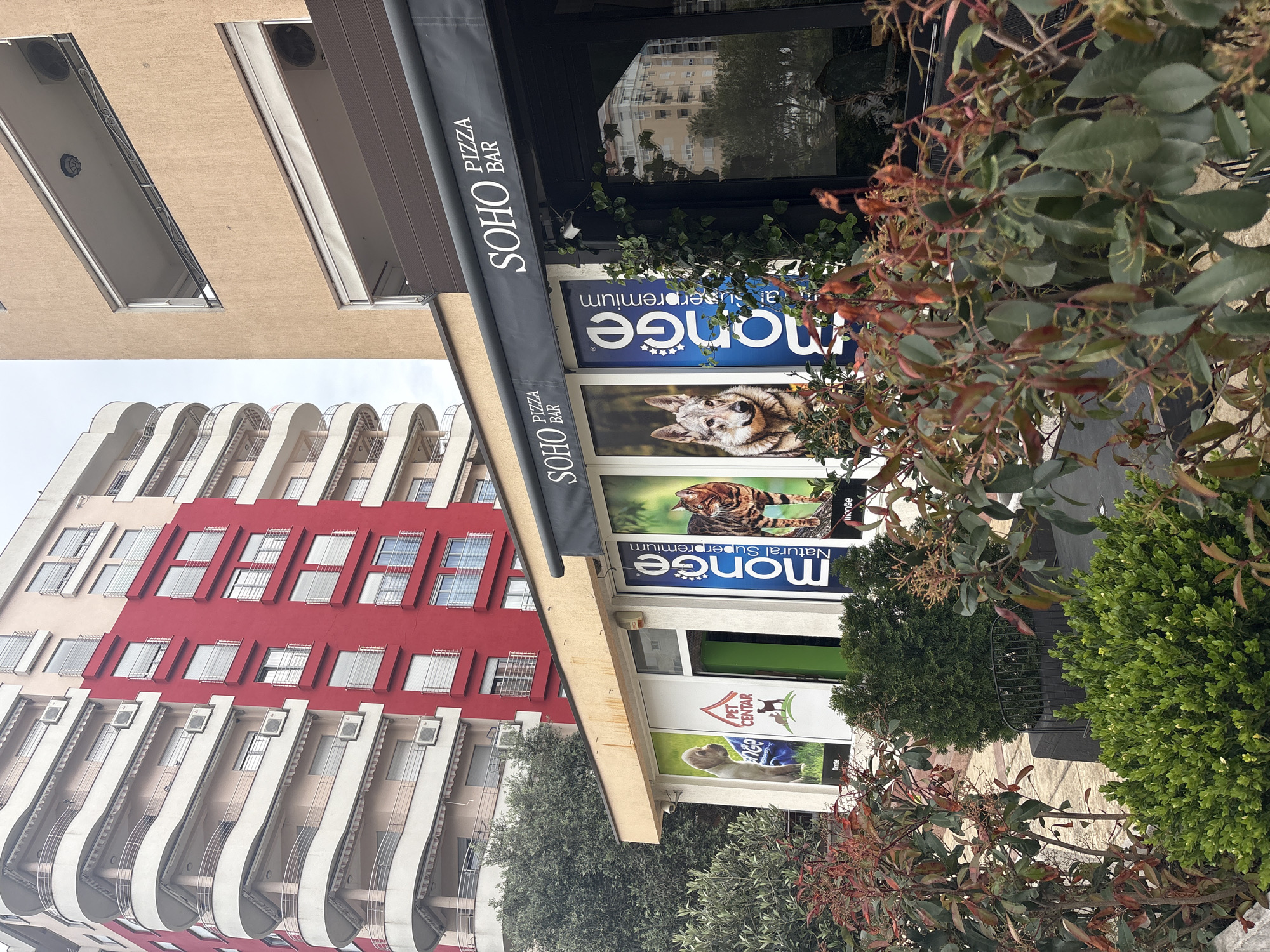 The image shows a commercial area in Bar, Montenegro. A building with a red and white facade and multiple balconies is visible in the background. The foreground features a business storefront with an awning labeled "SOHO PIZZA BAR". Below the awning are advertisements for "Monge Natural Superpremium" pet food, displayed with images of a dog and a cat. The entrance to "PET CENTAR" is also visible. Shrubbery is in the foreground, partially obscuring the view of the storefront.