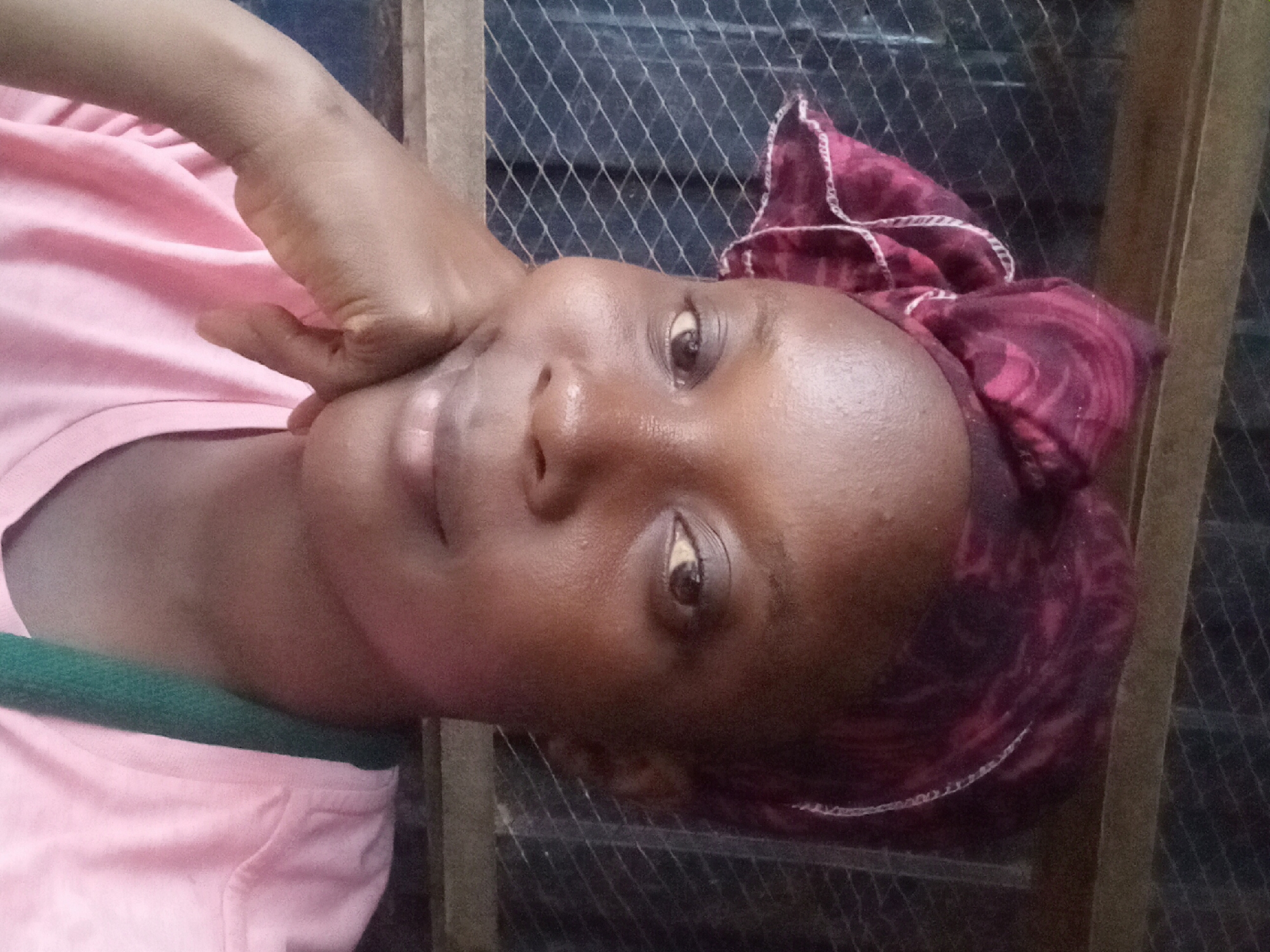 A close-up, rotated image shows a woman with dark skin resting her face on her hand. She is wearing a pink t-shirt with a green strap visible at the shoulder. She has a patterned burgundy and pink headscarf tied on her head. Behind her is a metal mesh screen with a wooden frame. The location is unspecified, and no evidence of Sabon Tasha, Nigeria, is present in the image.