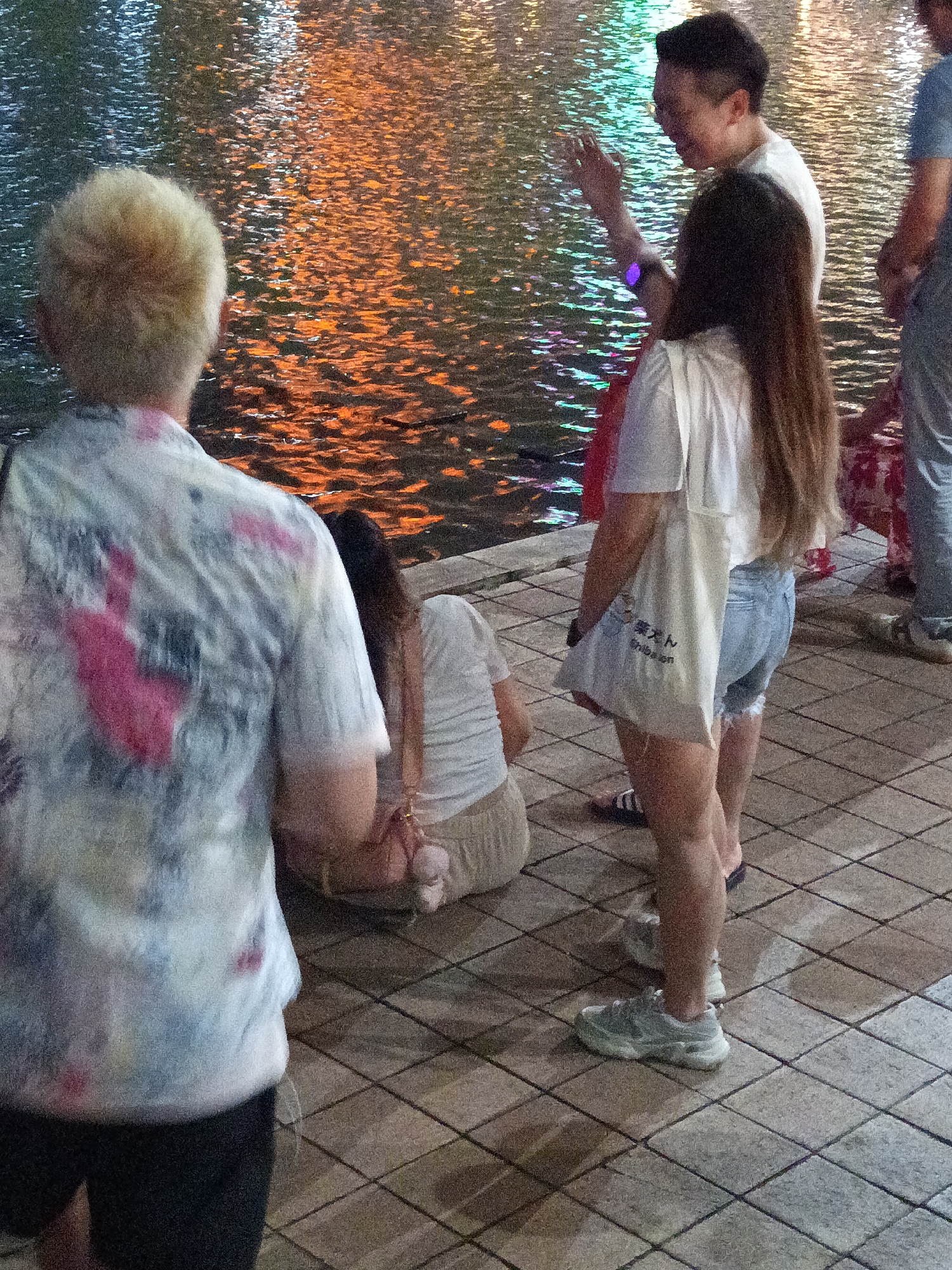 The image depicts a group of people standing by a body of water at night, likely in a public park or promenade area. The water reflects colorful lights, suggesting the presence of illuminated decorations or buildings nearby. A man in a white t-shirt is seen gesturing with his hand while talking to a woman with long brown hair who is wearing a white t-shirt and denim shorts, carrying a canvas tote bag. To the left, a person with short blonde hair is wearing a patterned shirt and dark shorts. In the background, another individual in a blue t-shirt and jeans is partially visible. The ground is paved with square tiles. The overall atmosphere appears casual, and the individuals seem engaged in conversation. The location is the central park of Kuala Lumpur, Malaysia.