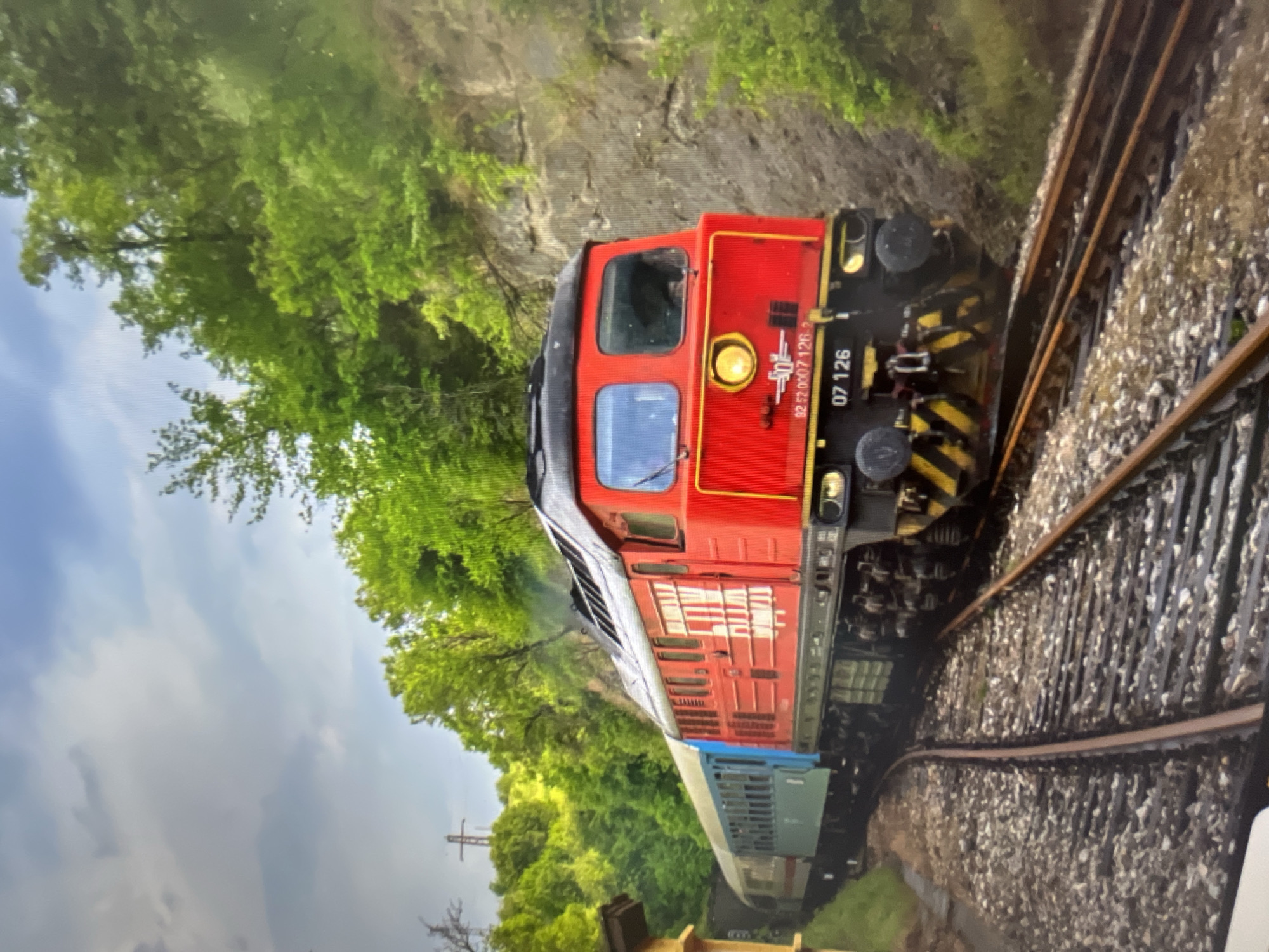A red and black diesel locomotive pulls a passenger train along a track through a wooded area. The train is moving from left to right, curving towards the viewer. The locomotive has a large yellow headlight and the number "07 126" on its front. The passenger cars behind the locomotive are a muted blue and white. The tracks are laid on a bed of gravel. The surrounding environment is lush and green with trees and rocky inclines. The sky is overcast with scattered clouds. There are no people visible in the image. The scene is likely a scenic route outside of a city, given the natural surroundings.