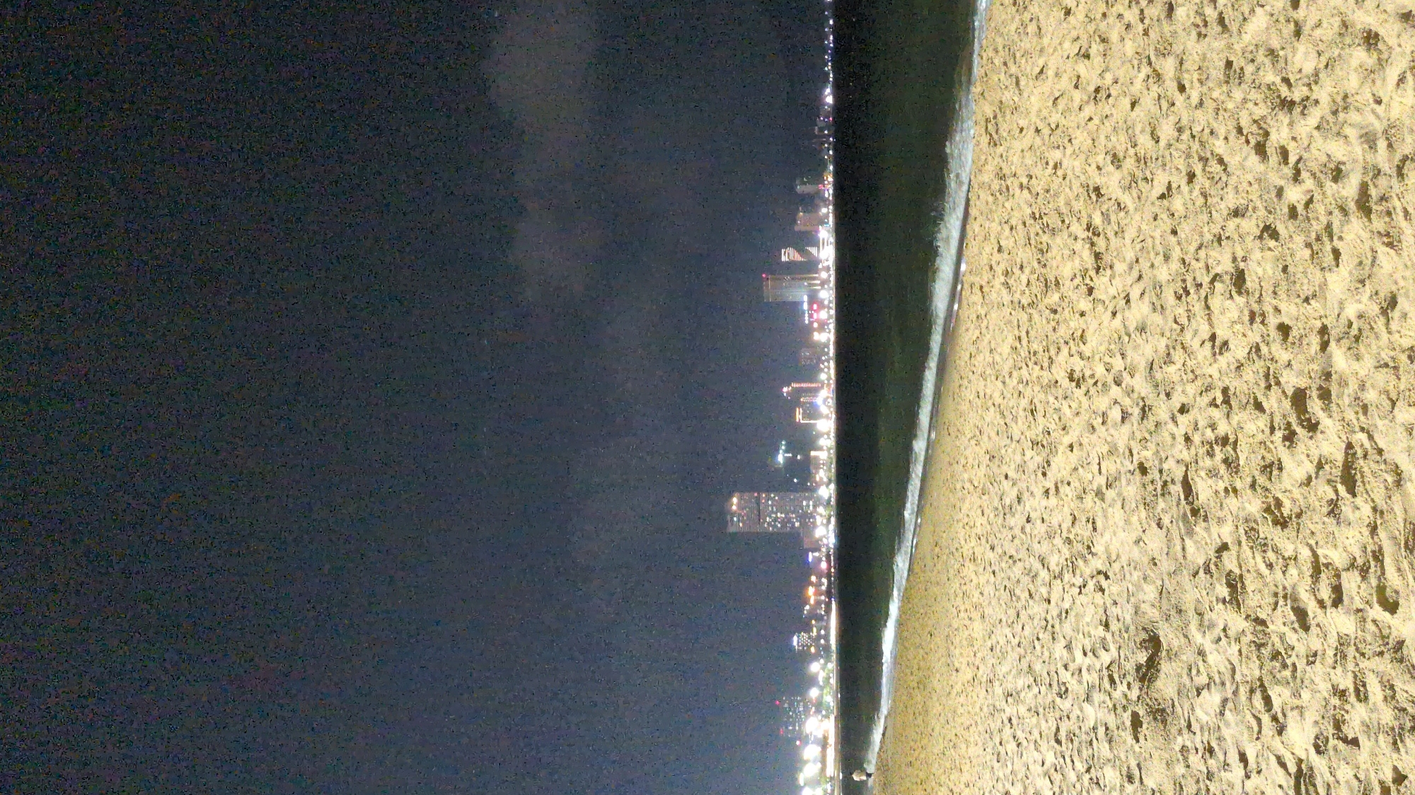 At night, the city of South Quy Nhon Ward, Vietnam is seen along the coastline. The city is dimly lit with lights from buildings and streetlights. The beach is in the foreground. The sky is dark.