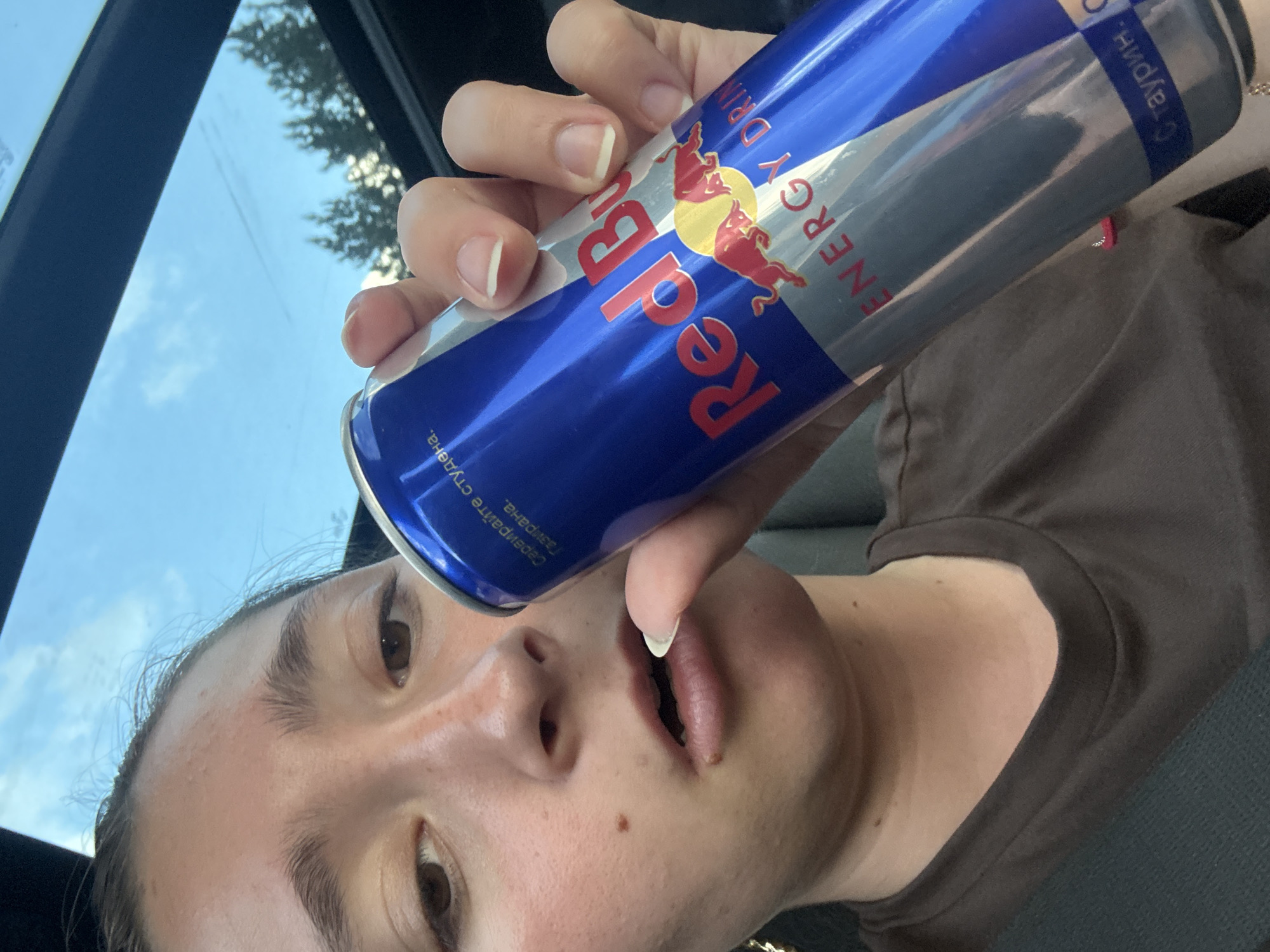 A close-up, selfie-style shot shows a young person's face and a hand holding a Red Bull energy drink can. The person is looking towards the camera with their mouth slightly open. The Red Bull can is predominantly blue and silver, with the brand's logo and text visible. The background suggests an outdoor setting, possibly a car interior with a sunroof showing the sky and trees. The image is not set in Dolno Varshilo, Bulgaria.